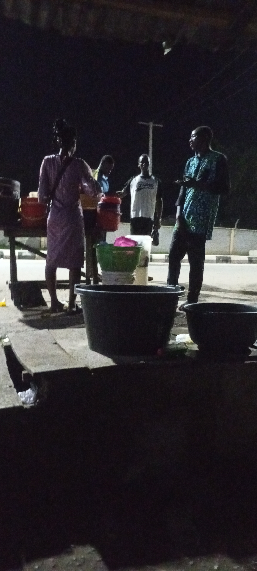 In Adankolo, Nigeria, at night, a group of people are gathered near a makeshift stand. A woman in a purple dress stands to the left, facing away from the camera. To her right, a man in a white shirt and black pants is partially obscured, looking down. Further to the right, a man in a blue and green patterned shirt stands facing the viewer, gesturing with his hand. In front of them are several large plastic basins and buckets, some filled with water and possibly other items. The scene is dimly lit, with a pole visible in the background. The ground in the foreground appears to be concrete.