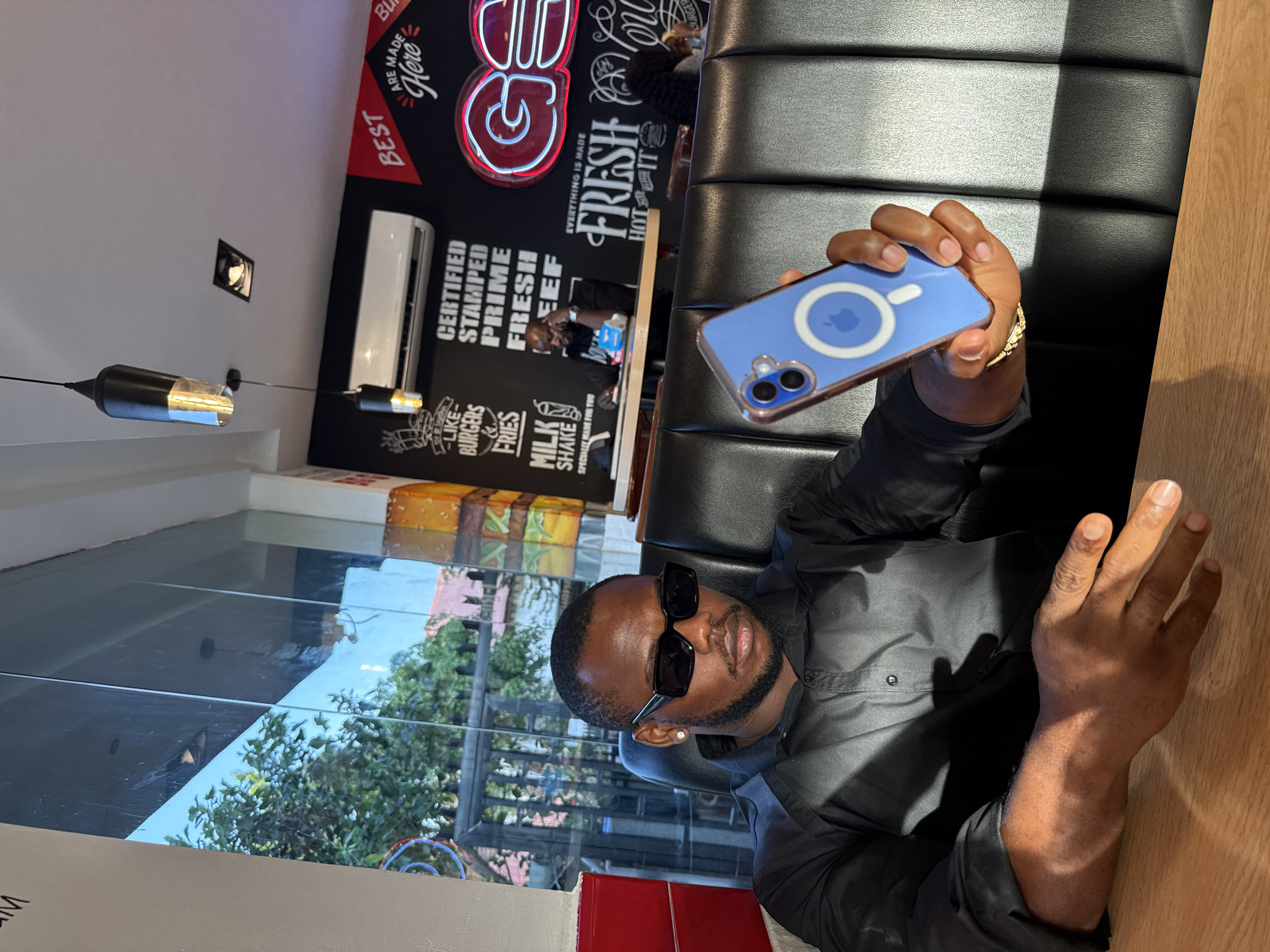 A man is holding a blue smartphone with a visible Apple logo. He wears sunglasses and a dark-colored shirt. The background shows a restaurant interior with menu boards advertising burgers, fries, and milkshakes. Text on the menu boards include "Certified", "Stamped", "Prime", and "Fresh". There is also a sign that says "Best are Made Here". The restaurant appears to be located in the city of Abuja, Nigeria.