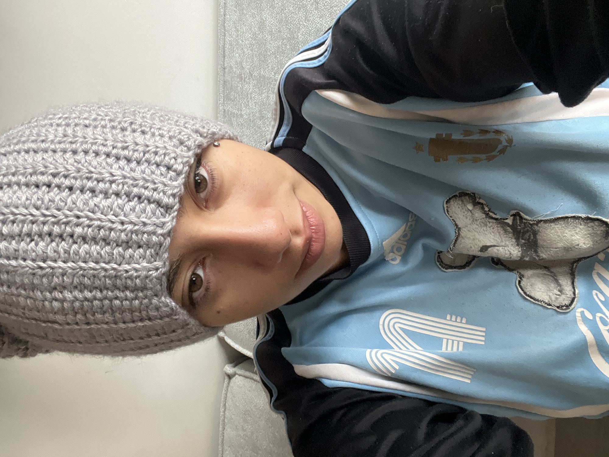 A person is wearing a gray crocheted beanie and an Argentina national team jersey. The jersey is light blue and white, with black sleeves and the Adidas logo. The person has an eyebrow piercing. The background appears to be a gray couch. The city and specific location within Buenos Aires, Argentina, cannot be determined from the image.
