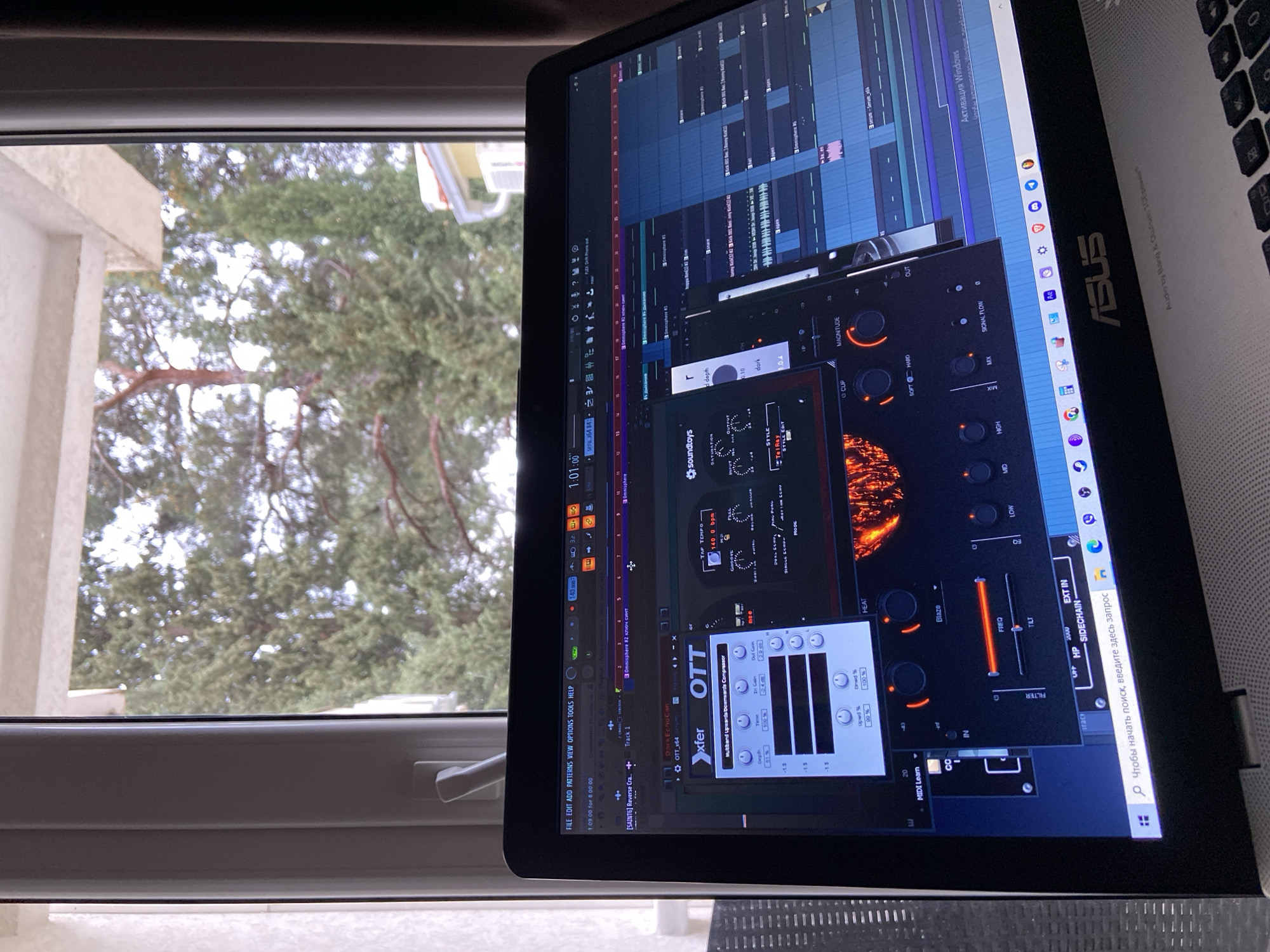 The image displays an indoor perspective through a window located in the city of Šušanj, Montenegro. In the immediate foreground, an open ASUS laptop with a black screen bezel is visible, resting on an unidentifiable light-colored surface. The laptop screen shows a Digital Audio Workstation (DAW) interface, consistent with FL Studio, displaying multiple audio tracks and two prominent plugins: Xfer OTT_x64, a multiband compressor, and a partially visible Soundtoys plugin with 'SATURATION' and 'Tel-Ray' style controls. The DAW's timeline indicates a timestamp of '1:01:00' and a tempo of '140 bpm'. The Windows taskbar and a Russian-language search bar are present at the bottom of the screen, along with a partial "Activate Windows" watermark. To the left and top-left, a white PVC window frame with a gray handle is visible. A dark, textured curtain or blind partially obscures the view on the right side of the window. Through the window, an outdoor scene is visible, dominated by numerous evergreen trees, including pines and cypress, under what appears to be an overcast or bright sky. In the lower right background, a section of a yellow building with white trim, a red-tiled roof, and an external wall-mounted unit is discernible. No individuals are present in the image.