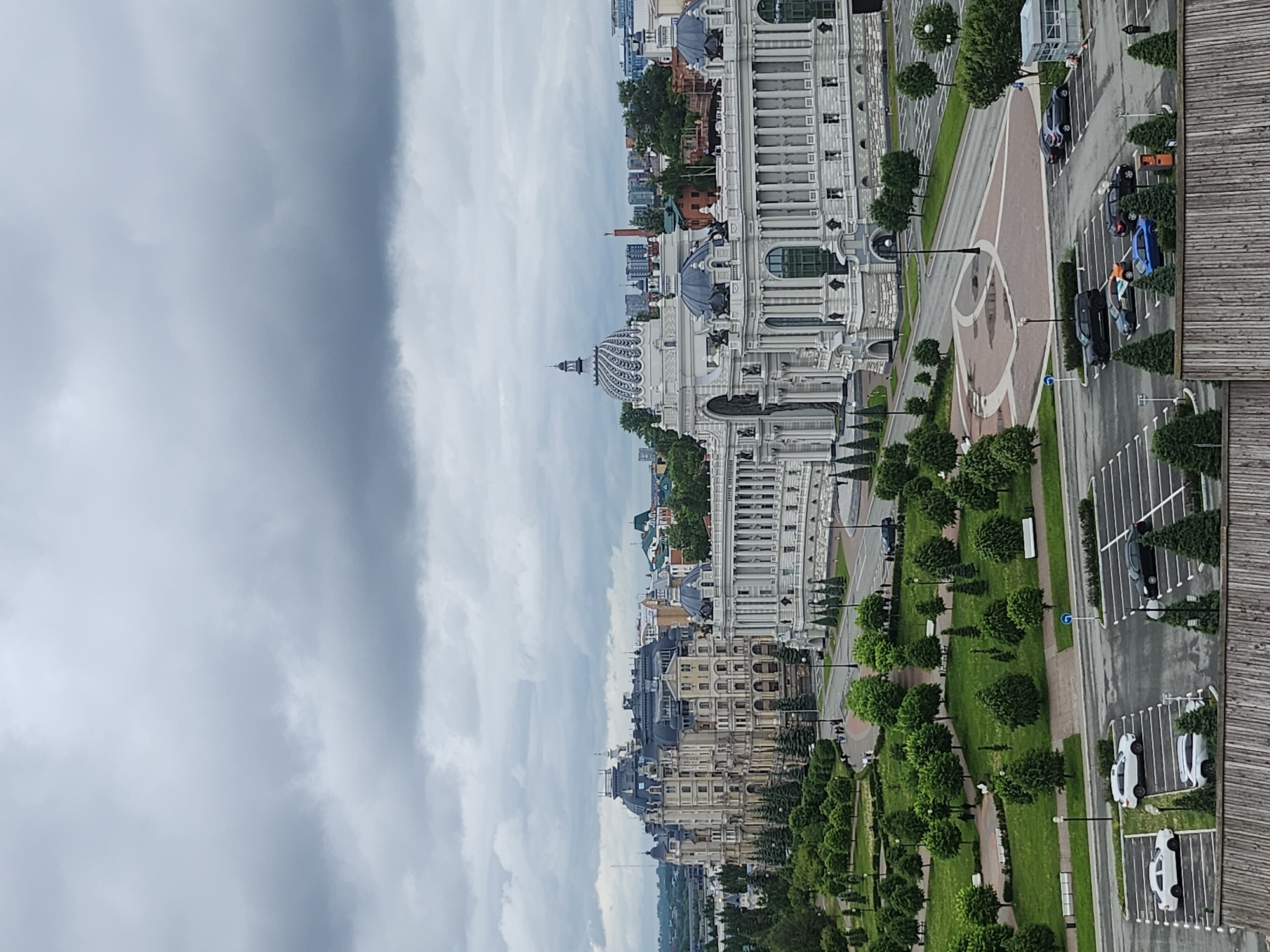 The image shows an aerial view of an urban landscape on a cloudy day. The architectural style is primarily European, with a notable white building that exhibits classical influences, including domes and ornate detailing. Below this main structure are other buildings with similar styles but in neutral tones, suggesting a historical or cultural district. A parking lot and road border the right side of the image. There are parked vehicles, streetlights, manicured greenery, and pedestrian walkways. The sky is overcast, indicating possible inclement weather.