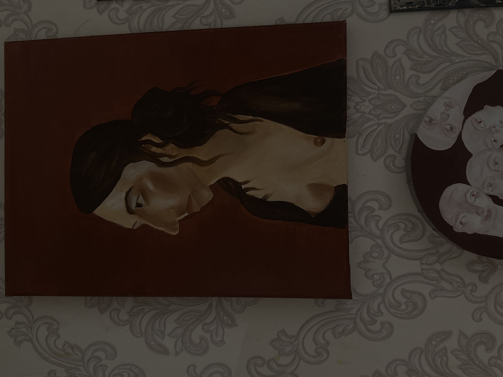 Two paintings are depicted on a wall. The left painting is a horizontal canvas with a dark red background. It features an oil painting of a person's profile, rendered in shades of brown and beige. The person has dark hair and is depicted with a somber or introspective expression. The right painting is a circular canvas, also with a dark background. It displays a cluster of stylized human faces painted in white with red accents around the eyes and lips. The faces are close together and overlapping, creating a somewhat abstract and perhaps unsettling effect. The wall itself has a light-colored damask wallpaper pattern. The overall scene suggests an indoor setting, possibly a home or an art display area. The lighting appears to be artificial and somewhat dim, casting shadows and contributing to a moody atmosphere. There is no visible text. The location is specified as ЖК «Борисоглебское», Russia.
