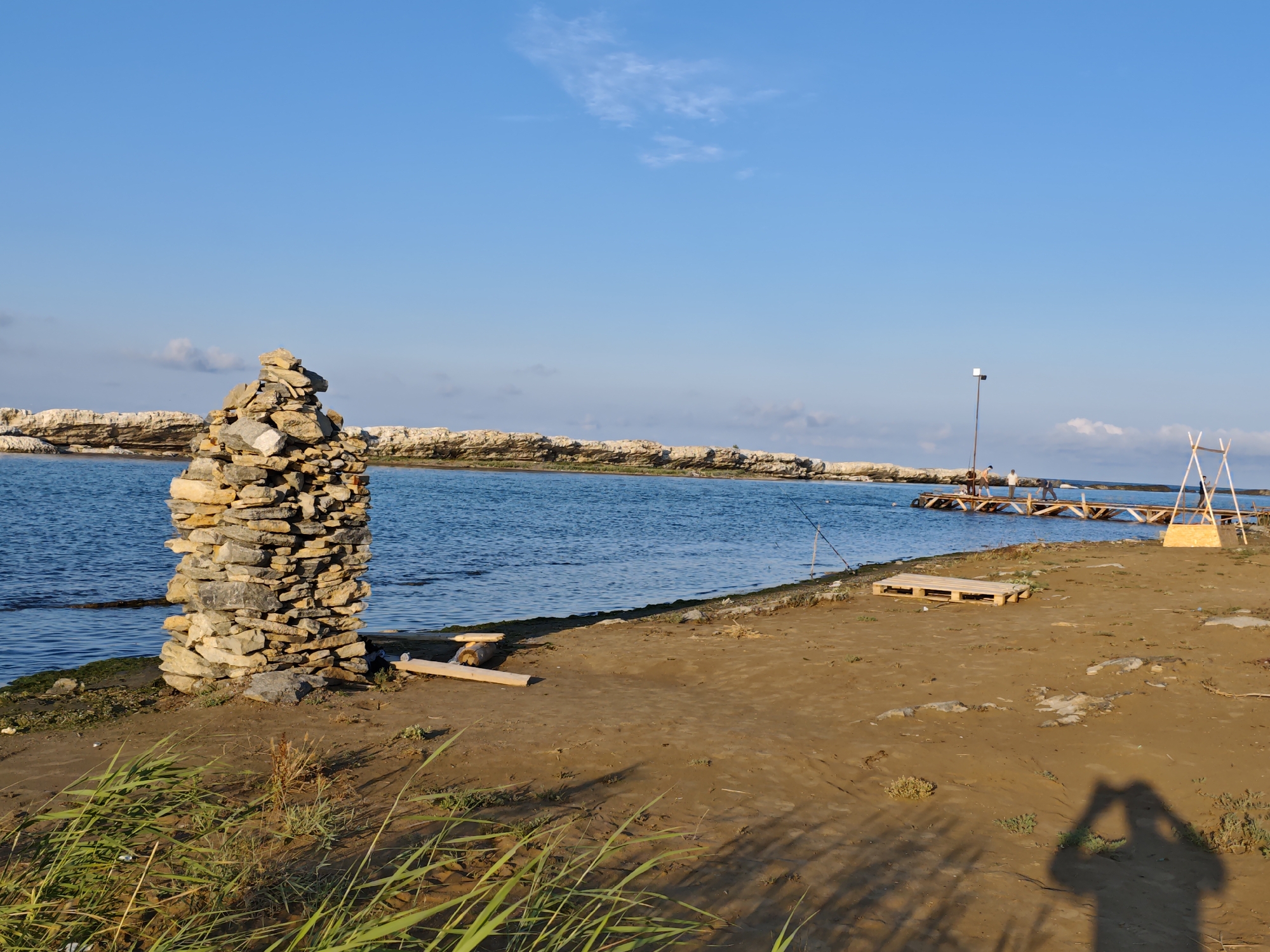 The image depicts a tranquil scene at a coastal location in Izberbash, Russia. In the foreground, a tall, irregularly stacked cairn of stones stands on a sandy shore. The water, a deep blue, stretches out to the horizon, meeting a rocky coastline. A wooden pier extends into the water, with several figures visible on it, some appearing to be fishing. A fishing rod is also planted in the sand near the shore. The sand is scattered with dry vegetation and some wooden planks. In the background, a clear blue sky is dotted with a few wispy clouds. The overall impression is one of a peaceful day by the sea.