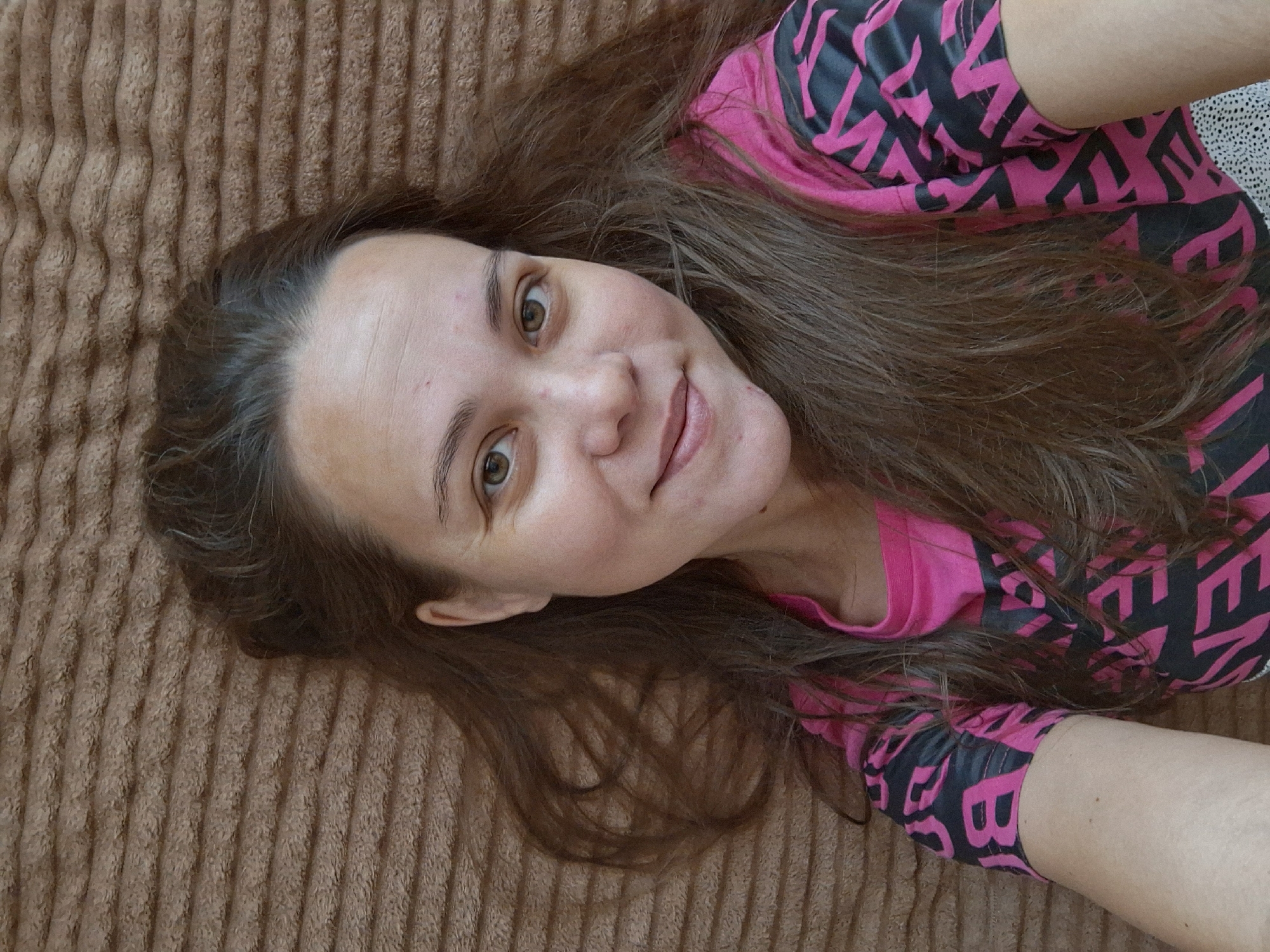 A close-up, overhead, sideways shot shows a woman lying on a textured brown blanket. She is wearing a bright pink shirt with black lettering that appears to be a repeating pattern of letters, possibly a brand name. Her head is tilted, and she is looking directly at the camera with a slight smile. Her brown hair is spread out around her head and shoulders. The lighting suggests it is daytime, and the indoor setting is indicated by the blanket. The overall mood of the image is casual and relaxed.