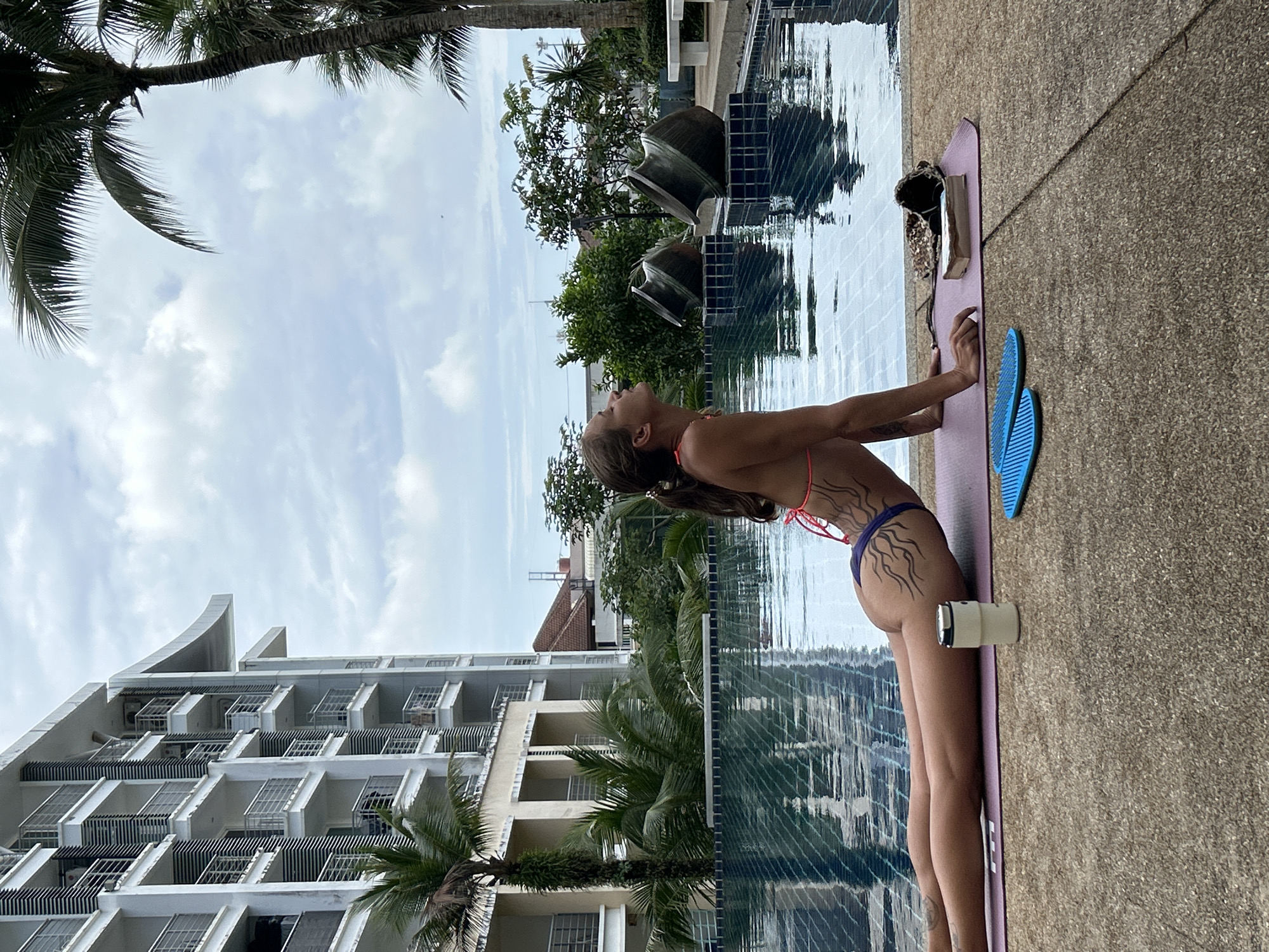 A young woman with brown hair tied in a ponytail is practicing yoga in a cobra pose on a purple yoga mat beside a swimming pool. She is wearing a bikini with a purple bottom and a thin orange string top. She has tribal-like tattoos on her back and buttocks. In front of her, on the mat, is a white insulated cup and two blue acupressure sandals. To the right of the mat, there is a small sequined bag and a book. The swimming pool is blue with tiled edges and reflects the cloudy sky and surrounding palm trees and a multi-story white building. The location is Kathu, Thailand, indicated by the tropical vegetation and architecture. The overall scene suggests a relaxed outdoor yoga session in a resort or residential complex.