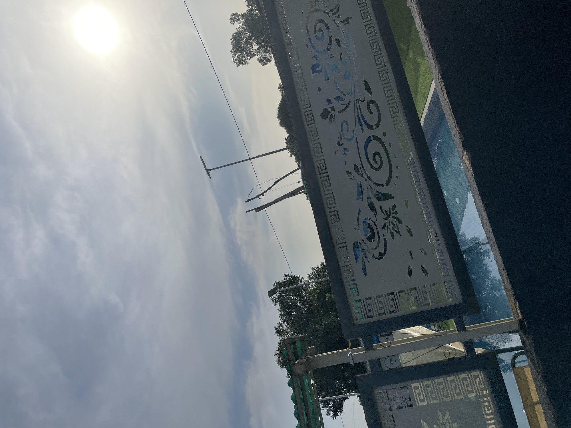 This image depicts an outdoor scene on a bright, sunny day in Jalingo, Nigeria. The sun is prominently visible in the upper left corner, casting a bright glare and suggesting it is mid-morning or afternoon. The sky is partly cloudy with streaks of white clouds against a lighter blue.
The foreground features a decorative metal screen with an intricate floral and geometric pattern, likely part of an architectural element like a fence or railing. Beyond this screen, glimpses of a swimming pool with clear blue water are visible, bordered by a green grassy area. This suggests the location is likely a residential area, a hotel, or a recreational facility.
In the background, several trees with dense foliage are scattered across the landscape, indicating a natural or semi-urban environment. Two tall utility poles with wires stretching across the sky are also visible, common features in many populated areas. The image is taken from a low angle, looking upwards towards the sky and across the scene. The overall mood is bright and serene, with no people or specific activities clearly depicted, focusing instead on the architecture and the natural elements of the setting.