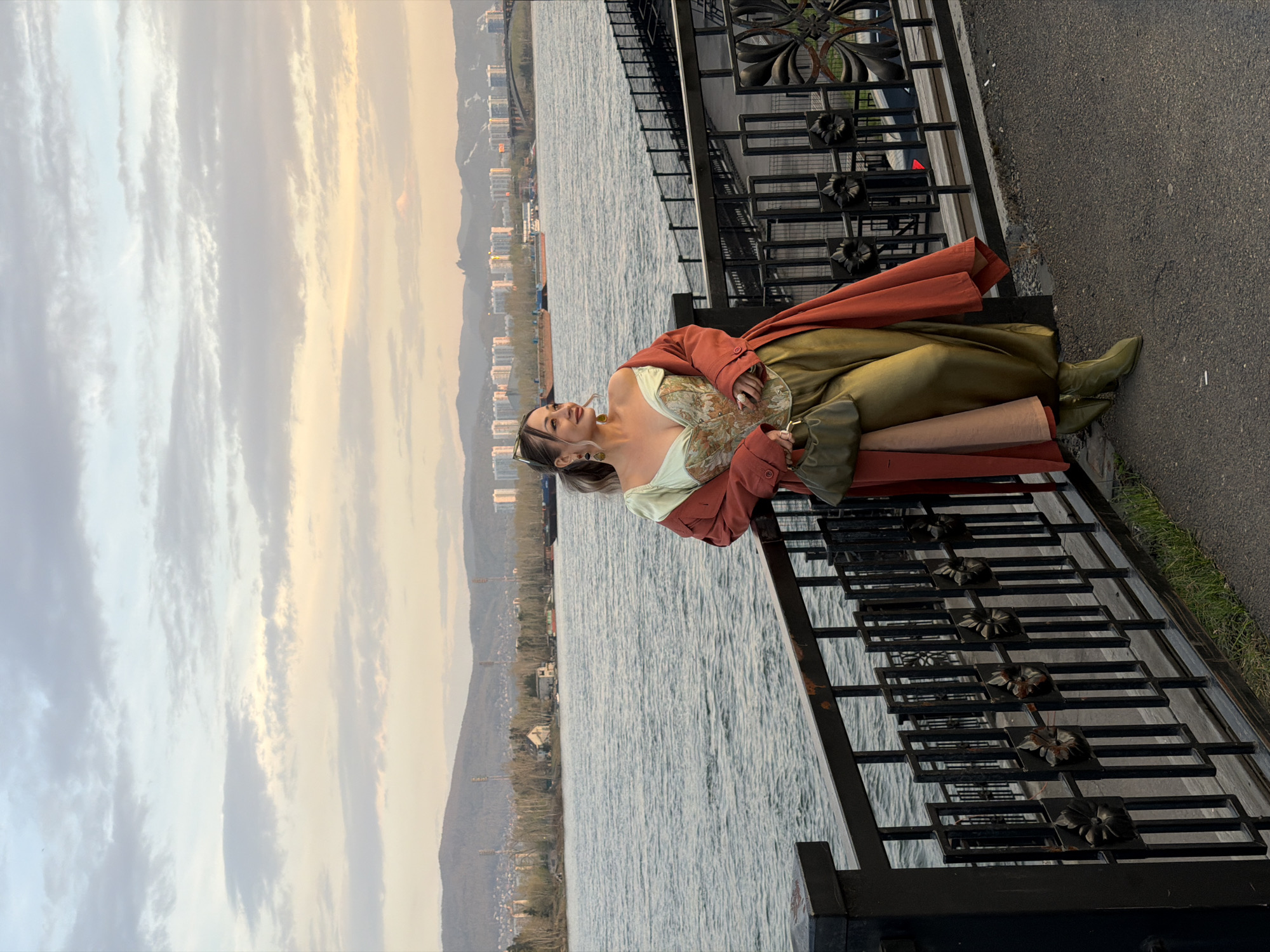 A woman is leaning back against a black metal railing on a paved walkway. She has long blonde hair and is wearing sunglasses, a light-colored floral-patterned top, a long olive-green skirt, and olive-green boots. A red coat is draped over her shoulders and arms. She is looking to the left.
Behind the railing is a large body of water. Beyond the water are buildings and a bridge, likely in Krasnoyarsk, Russia. Mountains are in the distance. The sky is cloudy with hints of yellow and blue. The scene is captured in a slightly tilted, landscape-oriented photograph. The railing is detailed with decorative floral designs.