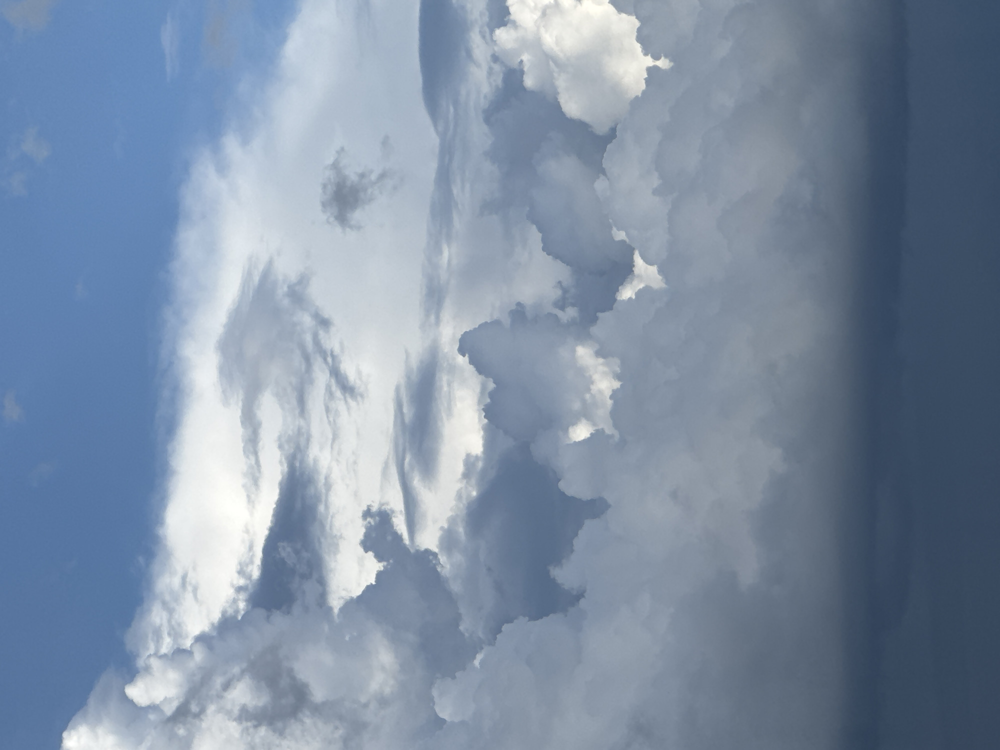 The image shows a section of the sky, likely taken from Krasnodar, Russia, given the prompt. The left portion of the frame reveals clear blue sky with a few scattered cumulus clouds. The majority of the frame is filled with larger, denser cloud formations. These clouds vary in color from bright white to dark grey, suggesting a variety of thicknesses and sun exposure. A heavy rain cloud is on the right. The image has been rotated 90 degrees clockwise.