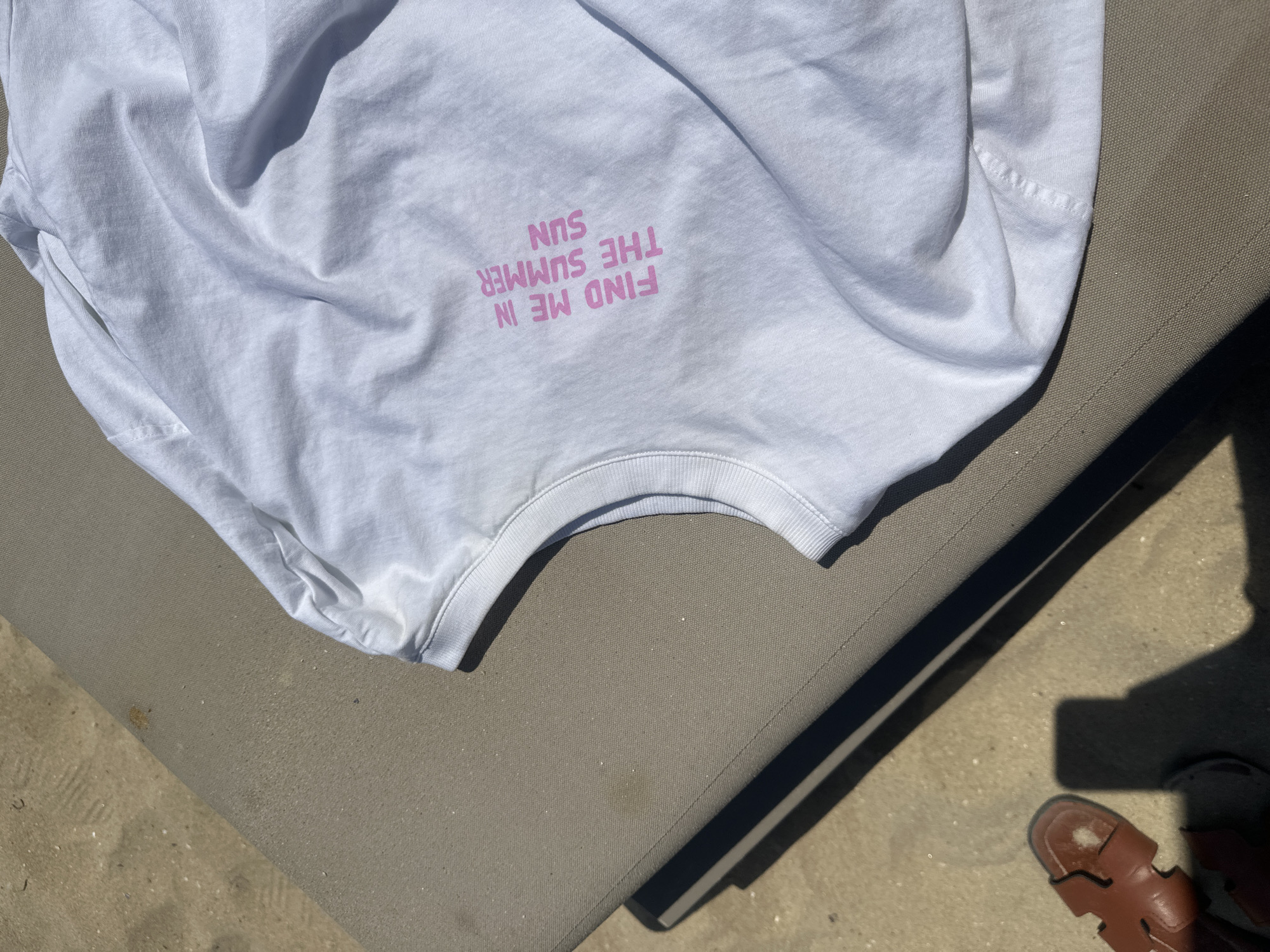 A white t-shirt with pink text is laid out on a lounge chair. The text reads "FIND ME IN THE SUMMER SUN". The lounge chair is on a sandy surface. A brown sandal is visible in the lower left corner of the image, casting a shadow. The scene appears to be outdoors, likely on a beach, in Nesebar, Bulgaria.
