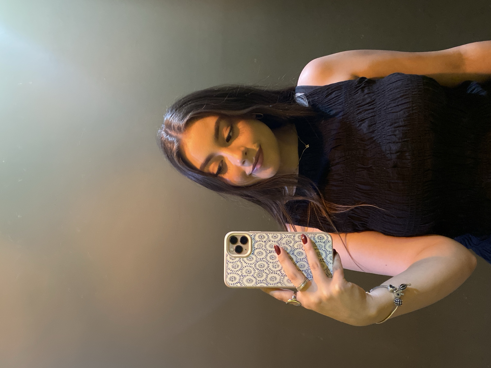 A young woman is holding a cell phone in her right hand, appearing to take a selfie. She has long, dark hair and is wearing a black top. She is also wearing a bracelet with charms and multiple rings on her fingers. The cell phone has a patterned case. The background is a solid, neutral color. The location is Unknown, Unknown.