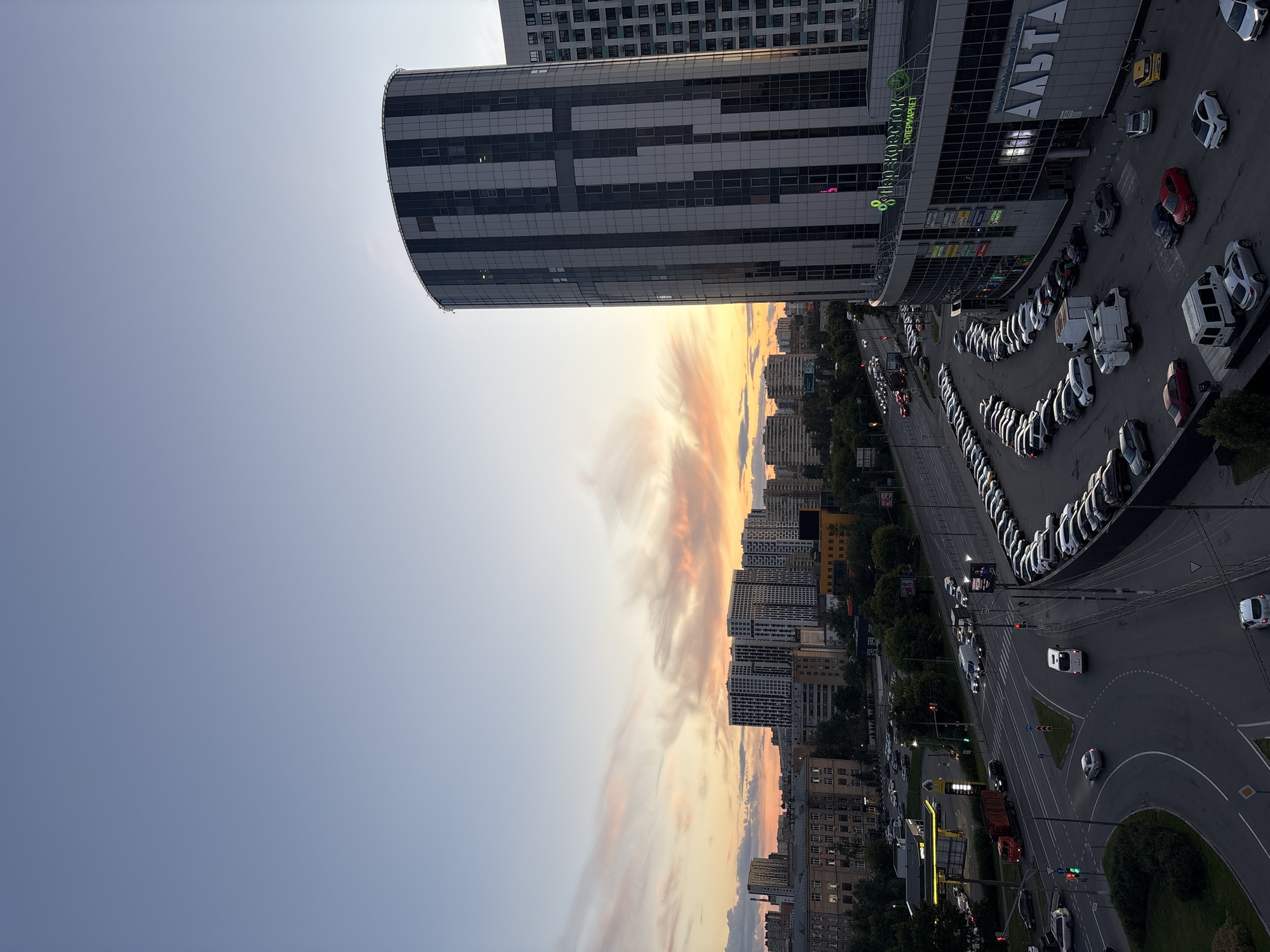 The image depicts a twilight cityscape in Korolyov, Russia. A large, modern, cylindrical building with a glass and dark paneled facade dominates the right side of the frame. Below it, a supermarket called "Перекрёсток" is visible. A vast parking lot filled with numerous cars is situated in front of the building, extending towards the left and curving around the road.
To the left of the cylindrical building, a multi-lane road is visible with traffic moving in both directions. A red truck and several other vehicles are seen on the road, along with traffic lights and pedestrian crossings. Further in the background, a dense collection of residential buildings and smaller commercial structures stretches across the horizon. The sky above is a gradient of blue, transitioning to shades of orange and pink, indicating a sunset with scattered clouds. The overall scene captures a typical urban environment during the evening, with a blend of modern architecture, infrastructure, and the natural beauty of the sky. No people are clearly discernible in the image, and no specific actions or interactions are evident.