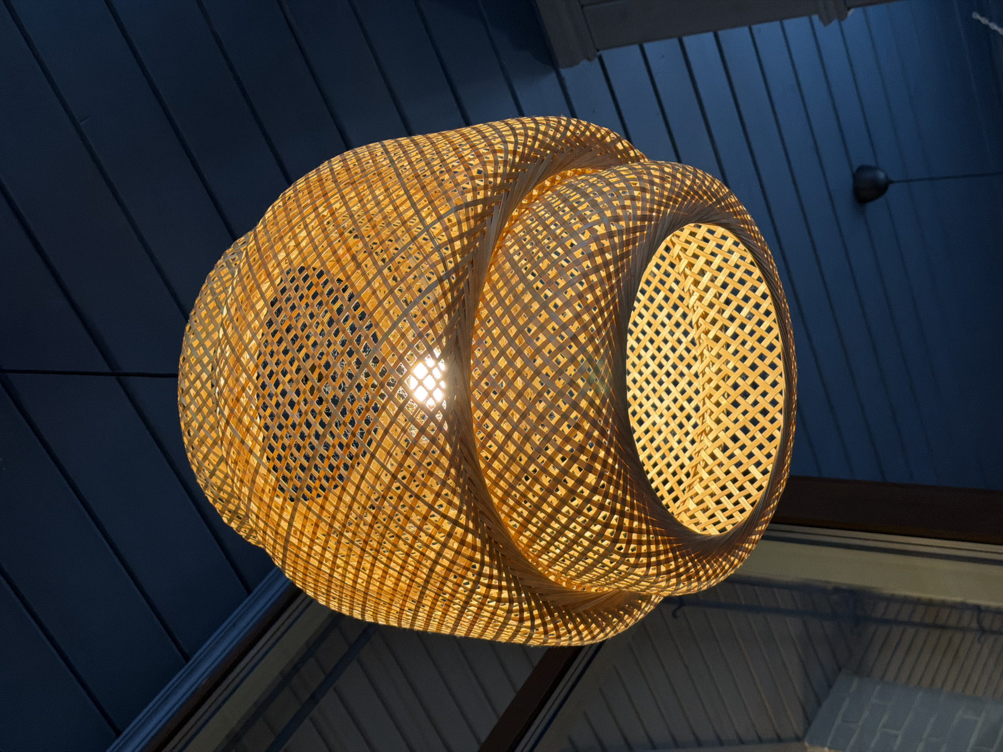 An illuminated, spherical, woven rattan lampshade hangs from a dark blue, slatted ceiling. The lampshade, with its intricate grid pattern, casts a warm, golden light. Through the open weave, a single light bulb is visible, emitting a bright glow. The background ceiling is painted a deep navy blue and appears to be made of wooden planks. On the right side of the frame, a white column with fluting is partially visible. To the left, a window reflects a brick-like structure and the outdoor environment. The overall scene suggests an indoor setting, possibly a restaurant or cafe, with a focus on decorative lighting. The image does not provide any information or clues about a specific location in the city of Krasnogorsk, Russia.