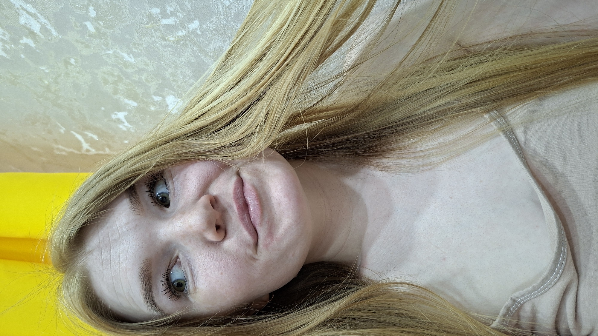 A medium-shot, close-up, rotated portrait features a fair-skinned woman with long, blonde hair, looking towards the top left of the frame. Her eyes are light, with visible eyelashes. She is wearing a light-colored top with a visible neckline. The background includes a yellow surface on the left side and a textured, light-colored wall behind her head. The location, Chaikovsky, Russia, is inferred from the context provided (although not directly visible in the image).