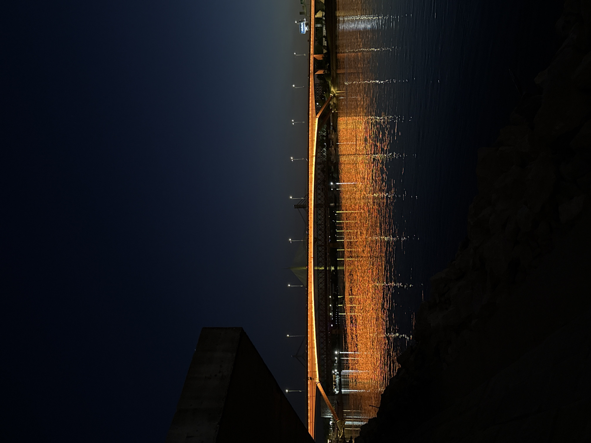 The image presents a striking nighttime view of the Ada Bridge spanning the Sava River in Belgrade, Serbia. The bridge horizontally dissects the frame, brilliantly illuminated with warm, golden lights that create a dramatic contrast against the dark surroundings.
The sky above is a deep, almost black, blue, indicative of late night. The architectural details of the cable-stayed bridge are visible, with its main structure silhouetted and its upper edge, cables, and numerous streetlights glowing brightly. These lights cast mesmerizing, elongated reflections of vibrant yellow and orange across the dark, still surface of the river below, creating a shimmering mosaic of light on the water.
In the foreground on the left, a concrete structure, possibly part of a riverbank or embankment, is partially visible. On the right, a dark, textured landmass or rocky shore frames the river. In the distant upper right, above the bridge, a faint blue sign with indistinct white text can be seen, though its content is unreadable due to the distance and low light. No people or specific activities are visible, emphasizing the serene and majestic presence of the illuminated bridge over the calm river. The overall scene conveys a sense of peaceful grandeur.