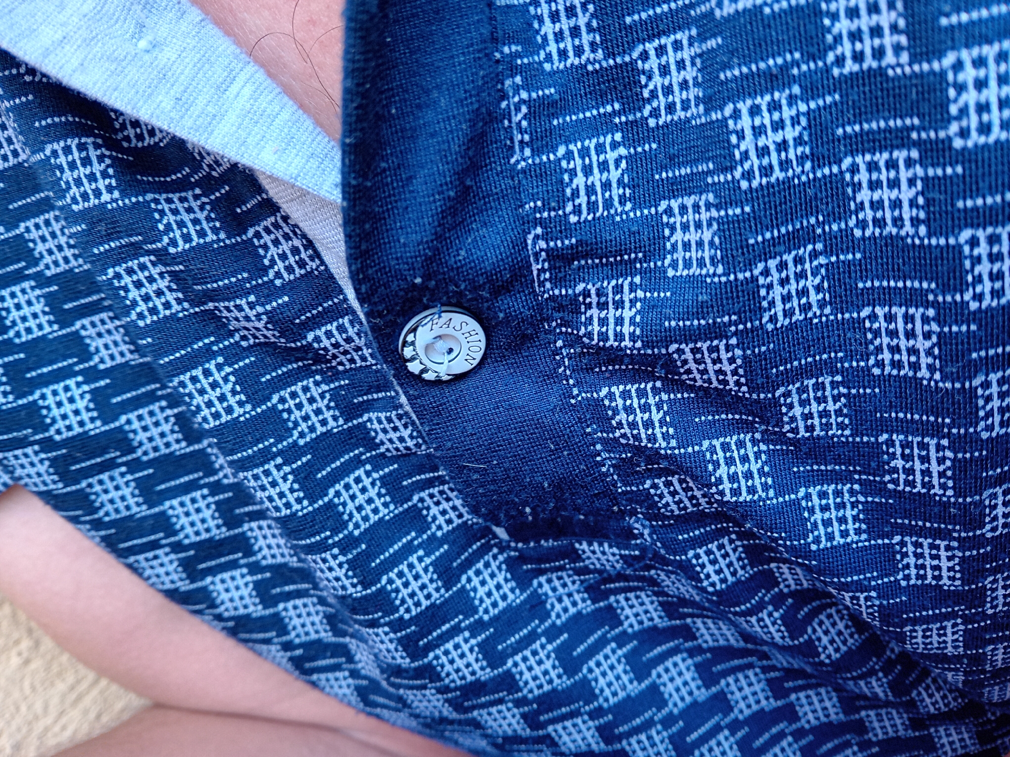 The image is a close-up, high-angle shot of a blue patterned shirt. The fabric of the shirt has a repeating white geometric pattern. A white button with the word "FASHION" is visible on the placket of the shirt, held in place by white thread. There is a glimpse of light-colored fabric underneath the blue shirt, possibly an undershirt. The image does not contain any information or context related to Zuberec, Slovakia.