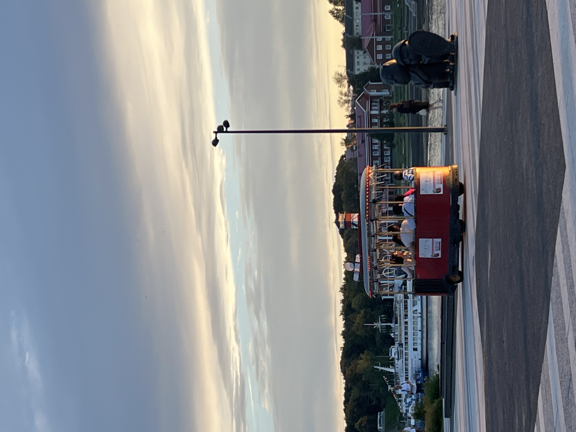 This image depicts a scene at the waterfront in Veliky Novgorod, Russia. In the foreground, a red open-air tourist trolley is moving on a paved pathway. Several passengers are visible inside the trolley, seated and looking around. To the right of the trolley, there are two dark-colored sculptures, appearing to be abstract figures. A lamppost with three spherical lights stands between the trolley and the sculptures. In the background, there are buildings with red roofs, lush green trees, and large ships docked on the water. The sky above is filled with clouds, with hints of sunset colors. A person in a dark outfit is walking on the pathway towards the sculptures. The overall atmosphere suggests a pleasant day for sightseeing.
