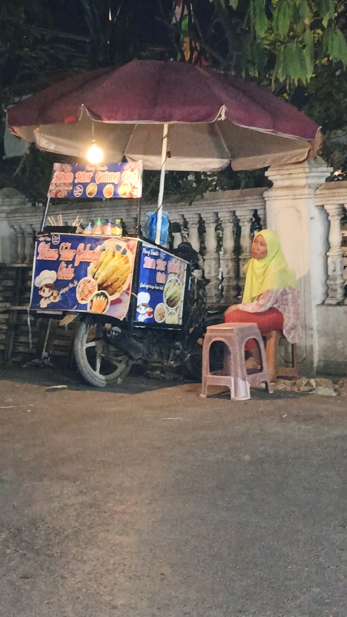 An evening scene in Muntur, Indonesia, depicts a woman vendor sitting on a stool beside her mobile food cart. The cart, adorned with colorful banners showcasing food items and cartoon chefs, is illuminated by a hanging light. The woman, wearing a yellow hijab and a patterned top, is seated on a pink plastic stool, looking to her right. Behind her, a stone wall with decorative elements and foliage suggests a semi-urban or residential setting. The food cart appears to be a motorcycle with a sidecar modified for street vending. The ground is paved asphalt, indicative of a street or road.