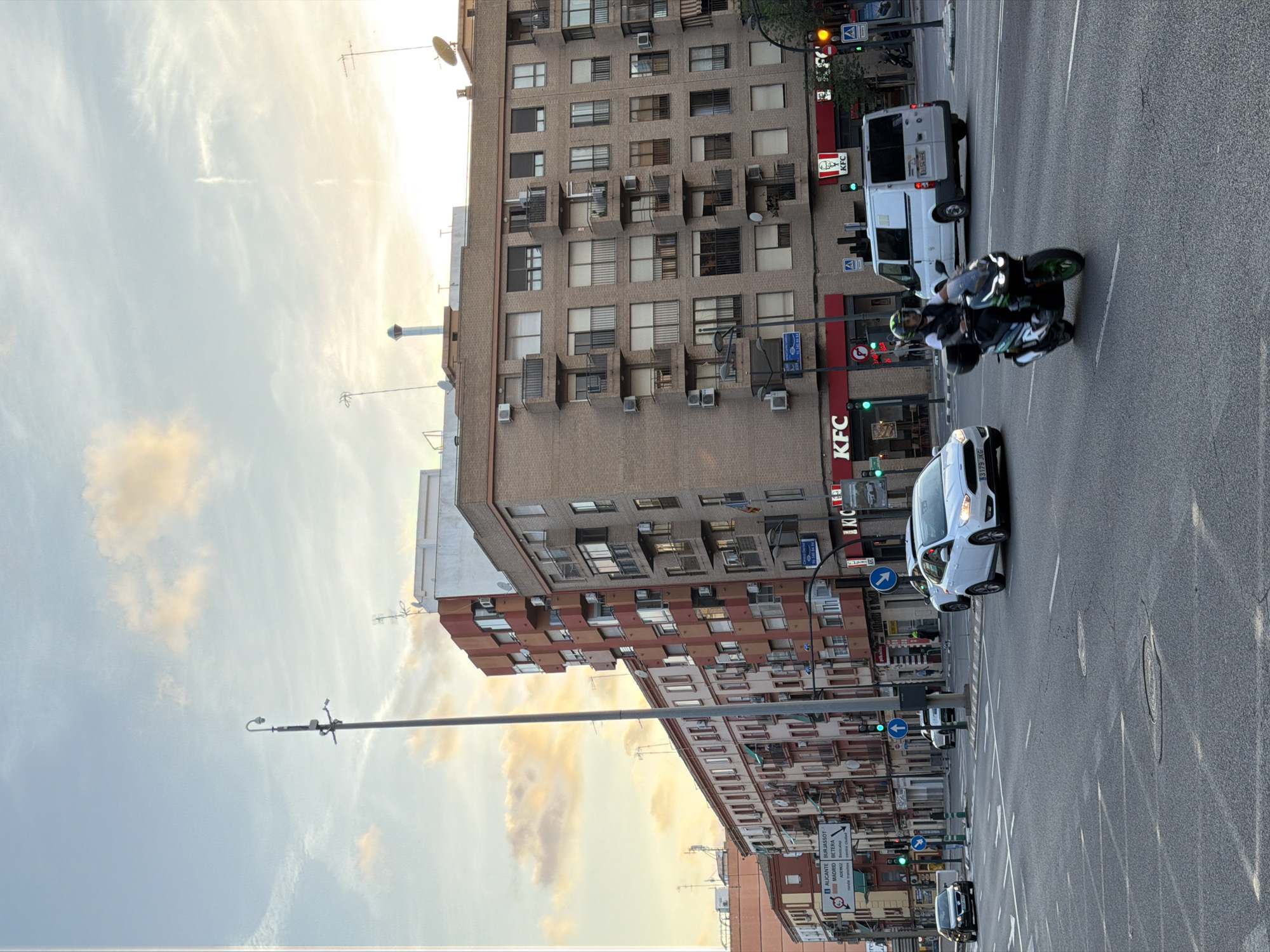 The image shows a street scene in an urban environment, likely in Spain given the road signs. The time of day appears to be late afternoon or early evening, with the sky displaying soft, warm colors from the setting sun and scattered clouds. The foreground is dominated by a road with lane markings, showing several vehicles in motion. A scooter with a rider is prominently featured in the middle of the frame, moving towards the right. To the left, a white car is driving away from the camera, and another white car is visible further down the road. A dark sedan is in the left lane, also moving away.
The background features several multi-story buildings lining the street. These buildings have a mix of architectural styles, with some featuring red brick facades and others painted in lighter colors. Many windows and balconies are visible. A KFC restaurant with its distinctive red signage is clearly visible on the ground floor of one of the buildings. Traffic lights and street lamps are also present, indicating an active intersection. Road signs are visible, including directional signs indicating cities like Alicante, Madrid, and Burjassot. The overall impression is a busy city street during a peaceful transition from day to night.