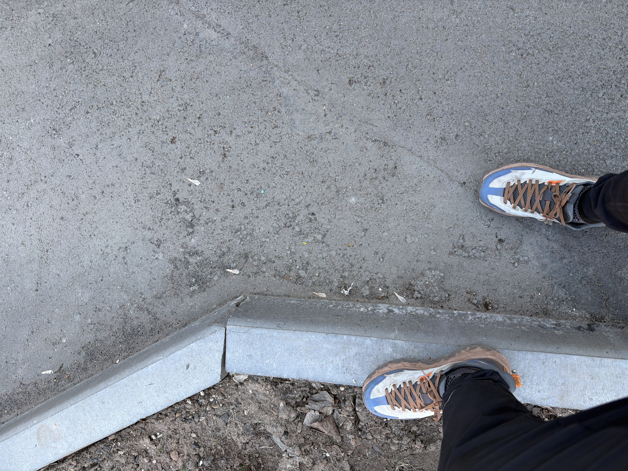 A street-level shot shows a section of sidewalk in Izhevsk, Russia. The sidewalk is made of gray concrete with a rough surface texture. Along the edge of the sidewalk, there is a raised concrete curb. A pair of feet are positioned on the sidewalk and adjacent to the curb. The feet are wearing running shoes with blue and white uppers and brown laces. The legs are covered in black pants. Patches of soil and sparse vegetation are visible beyond the curb.