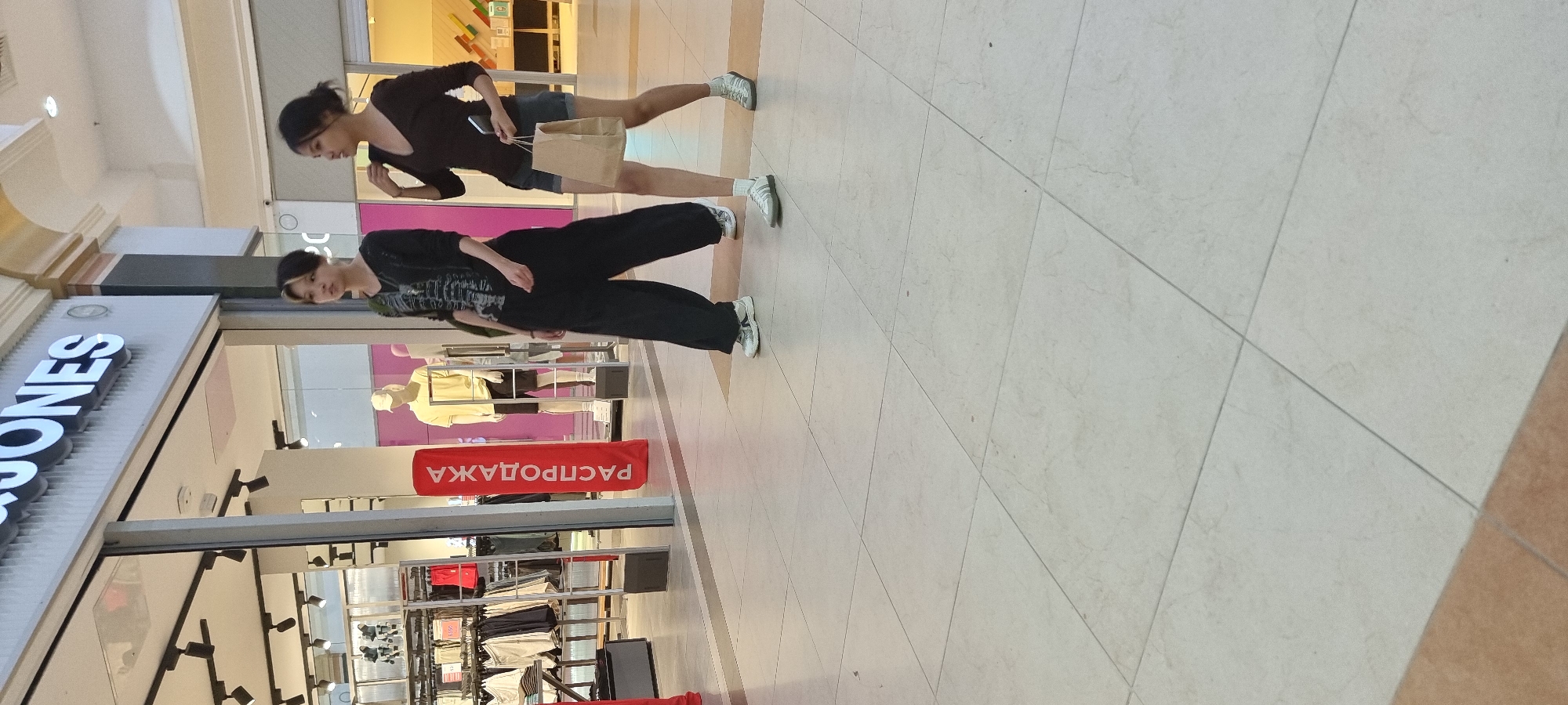 The image depicts two young women walking in a brightly lit indoor shopping mall. In the background, a storefront displays a "JACK & JONES" logo, suggesting the location is likely a branch of this clothing store. To the left of the storefront, racks of clothing are visible, along with mannequins. A large red banner with Russian text, "РАСПРОДАЖА" (meaning "SALE"), is prominently displayed in the foreground. Both women are dressed casually and appear to be shopping. The floor is tiled with a light-colored, square pattern. The scene suggests a typical retail environment within a city. Based on information associated with the store "JACK & JONES", this image was taken in the city of Aktobe, Kazakhstan.