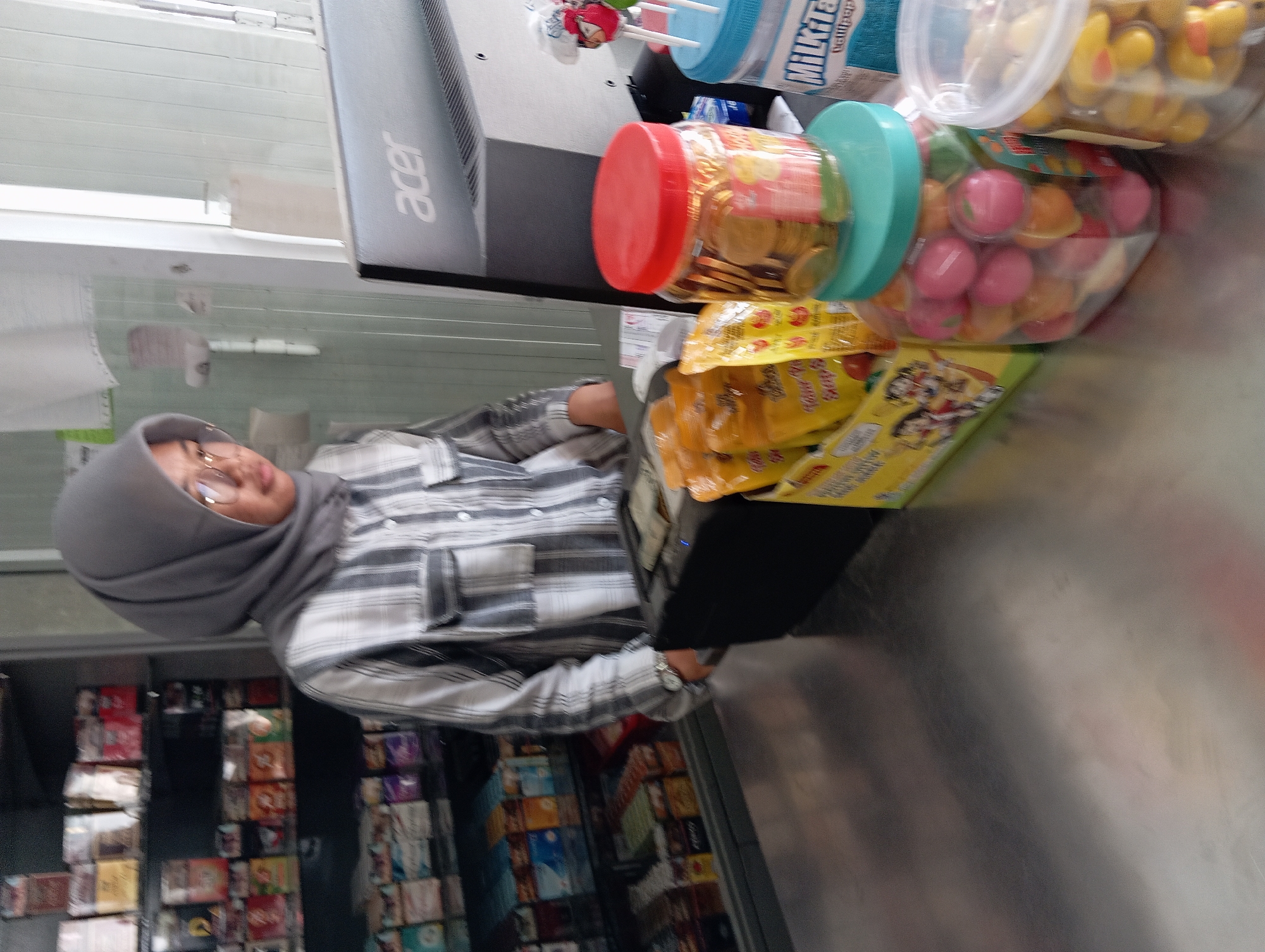 In the city of Srumbung, Indonesia, a young woman wearing a gray hijab and glasses stands behind a counter in what appears to be a convenience store or small shop. She is wearing a black and white striped button-up shirt. In front of her, on the counter, are various items, including a cash register, several bags of snacks, and jars filled with colorful candies. To her right, a computer monitor with the Acer logo is visible. Behind the woman, shelves are stocked with numerous products, including what appear to be cigarette packs. The environment is brightly lit, and the scene suggests a commercial transaction or inventory management.
