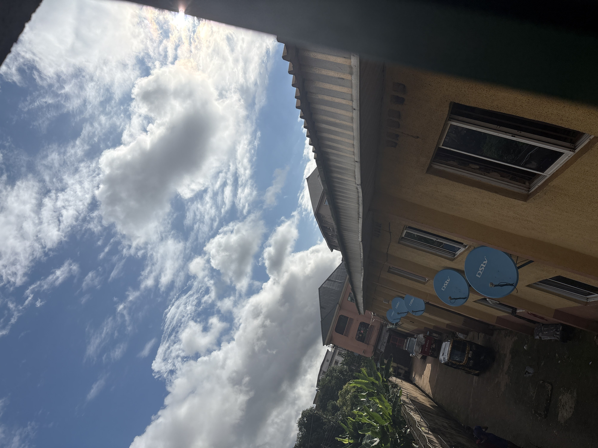 The image depicts a view from a building's exterior, looking upwards and across a narrow street in Onitsha, Nigeria. The sky is a bright blue with scattered, fluffy white clouds, illuminated by sunlight. In the foreground, a corrugated metal roof extends across the top portion of the frame, casting shadows. Below and to the left, a yellow building with multiple windows is visible. Several blue satellite dishes, marked with "DSTV," are mounted on the side of this building. Further down the street, a narrow alleyway is visible, flanked by buildings and vegetation on the left. Several vehicles, including what appears to be a three-wheeled auto-rickshaw and a small truck, are parked or moving along the street. The overall scene suggests an urban environment in Nigeria, characterized by multi-story buildings, visible infrastructure like satellite dishes, and typical street vehicles.