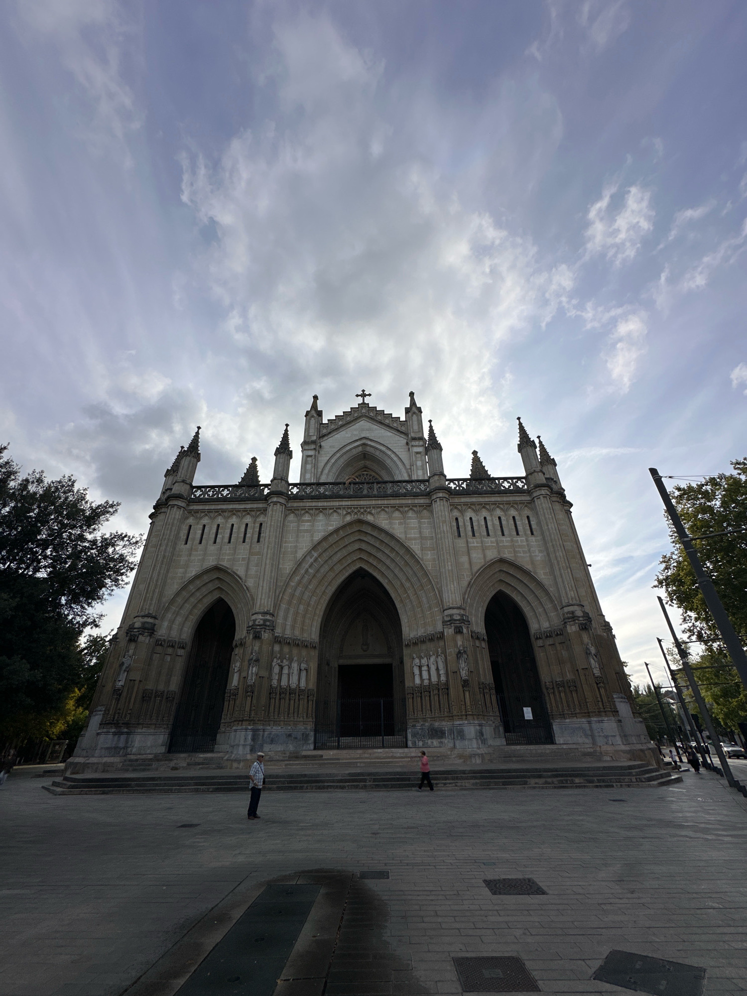 The image shows a wide, low-angle shot of the Vitoria-Gasteiz Cathedral, also known as the Cathedral of Mary Immaculate, located in Vitoria-Gasteiz, Spain. The cathedral's facade is made of stone and features Gothic architectural elements, including pointed arches, spires, and intricate carvings. The main entrance is marked by a large set of double doors.
In the foreground, the paved plaza in front of the cathedral is visible, with some people scattered about. To the left, a man in a light-colored shirt and hat stands facing the cathedral. To the right, a person in a pink shirt walks away from the cathedral. The sky above is filled with clouds, some of which are wispy and streaked, while others are darker and more dense. Trees can be seen on the left and right edges of the frame, and in the distance on the right, there are power lines and what appears to be a street with some traffic. The overall lighting suggests it is daytime, possibly late afternoon or early morning, given the soft light and shadows.