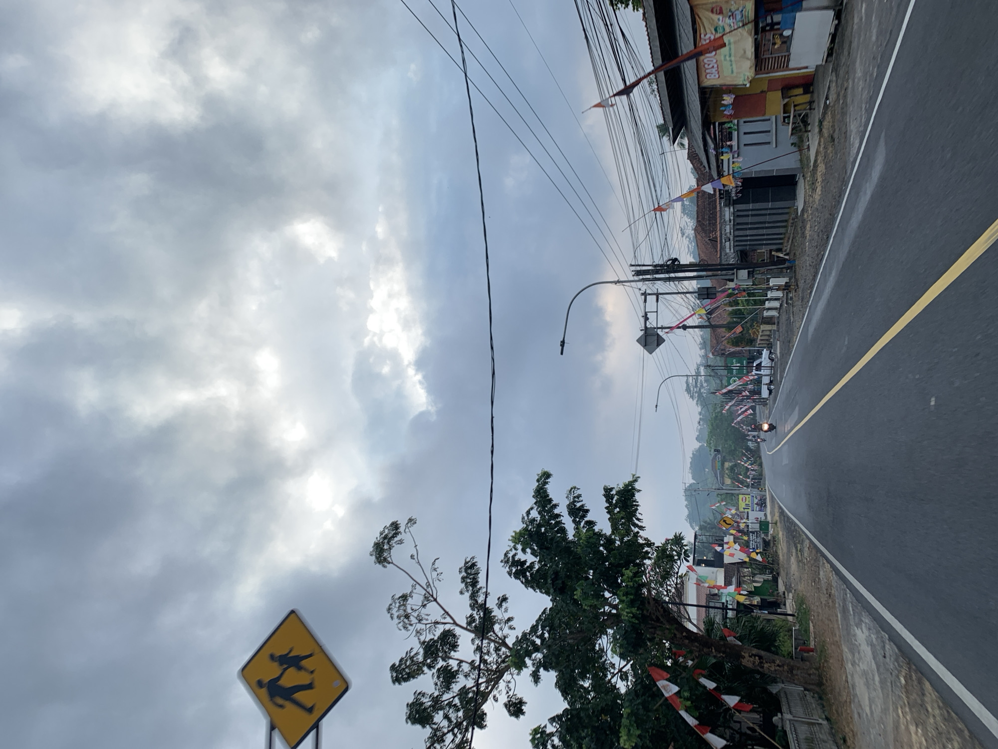 The image depicts a street scene in Cijeungjing, Indonesia, under a cloudy sky. A yellow diamond-shaped traffic sign with a silhouette of an adult and child walking is visible in the upper left quadrant. The street is paved with asphalt and features a yellow dividing line. Along the left side of the road, a large tree with dark green foliage stands, decorated with red and white triangular flags. Further down the street, houses and small businesses line the right side, also adorned with various colorful flags. Power lines stretch across the scene above the buildings. A motorcycle is being ridden down the road, and a white vehicle is parked on the left. The overall environment suggests a residential or commercial area in a town or village.