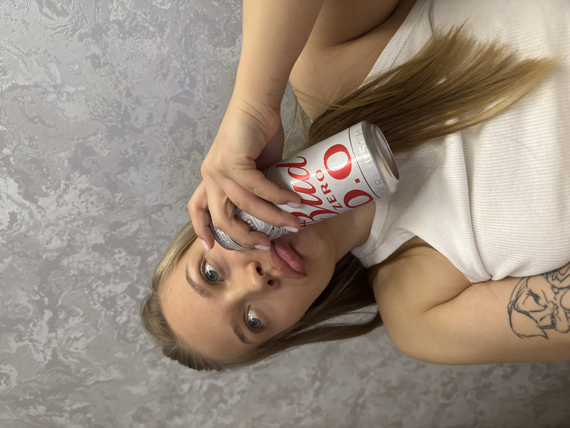 A young, fair-skinned woman lies on a gray and white patterned surface, presumably fabric or wallpaper. She is positioned with her head tilted to the left side of the frame. She is wearing a white top. Her blonde hair is partially visible. She holds a white can labeled "0.0" with red lettering in front of her face. She has light blue eyes and her lips are slightly puckered. There is a tattoo on her upper right arm. The location is unknown and there is nothing in the image to connect it to the city of Chaikovsky, Russia.