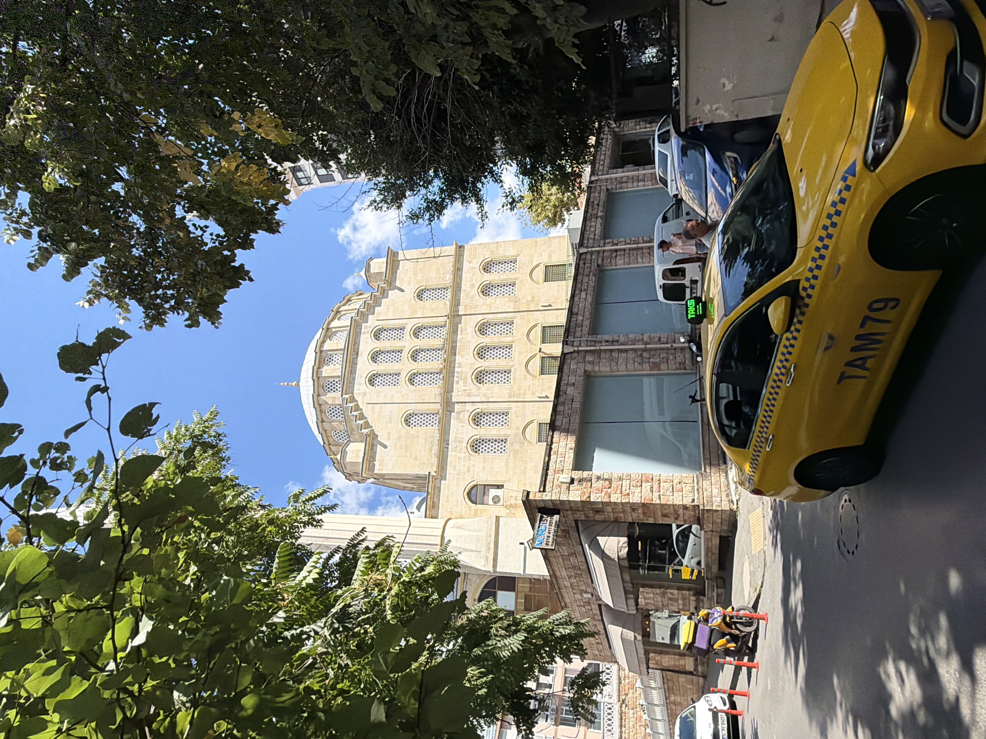 In Kadıköy, Turkey, a yellow taxi with the number TAM79 is parked on a street. In the background, a light-colored stone mosque with a dome and minaret is visible. Trees with green leaves frame the top of the image. The sky is blue with some clouds. Other vehicles, including a scooter, are also present on the street. A building with glass windows is situated in front of the mosque.