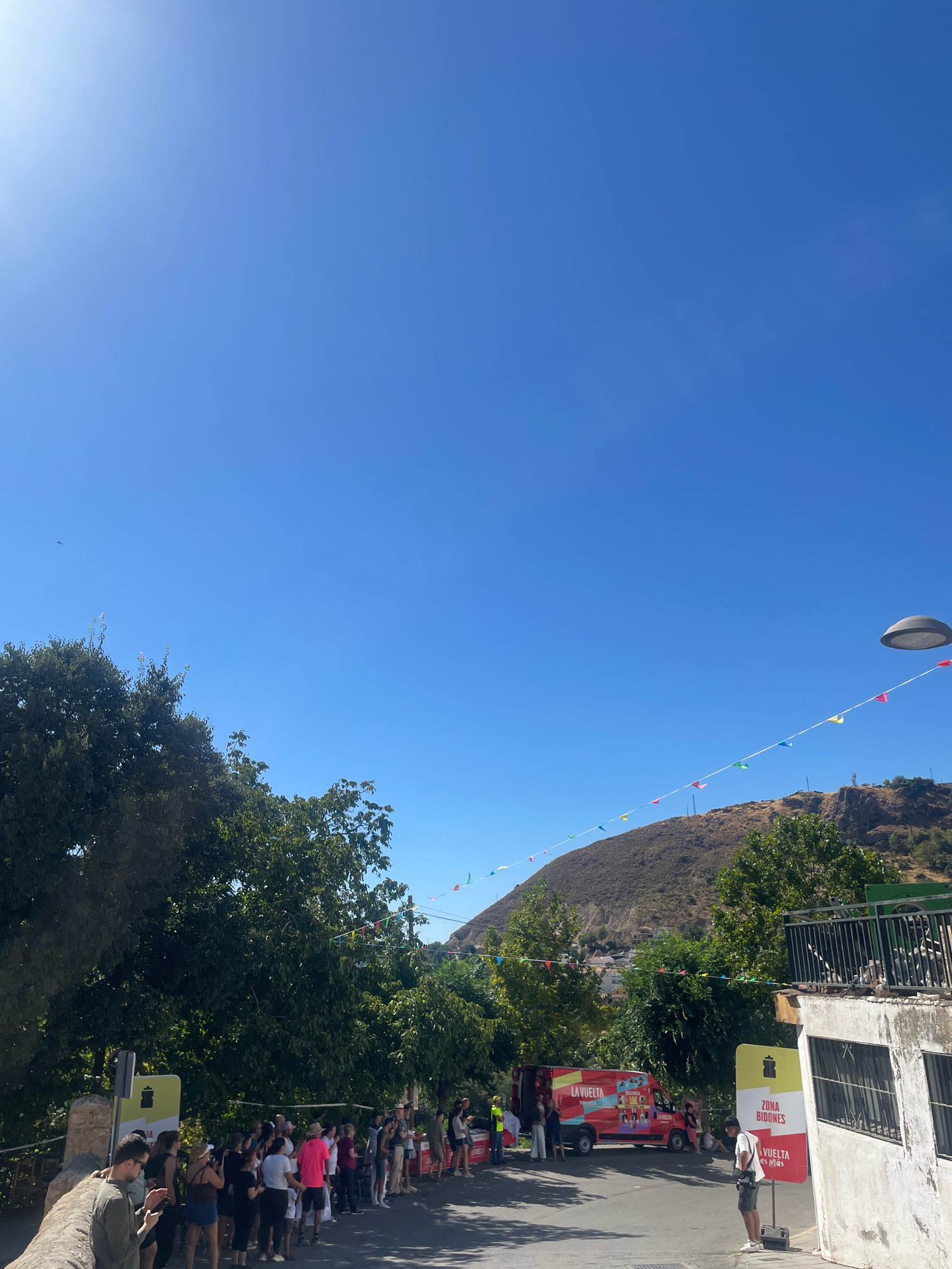 A group of people are gathered on a street in Güéjar Sierra, Spain, beneath a clear blue sky. A red van with "LA VUELTA" branding is parked on the street. Several trees line the background, and a hillside with sparse vegetation rises behind them. Colorful triangular flags are strung across the scene, and a lamppost is visible in the upper right corner. People are standing and observing, some are clapping. A sign reads "ZONA BIDONES" and "LA VUELTA".