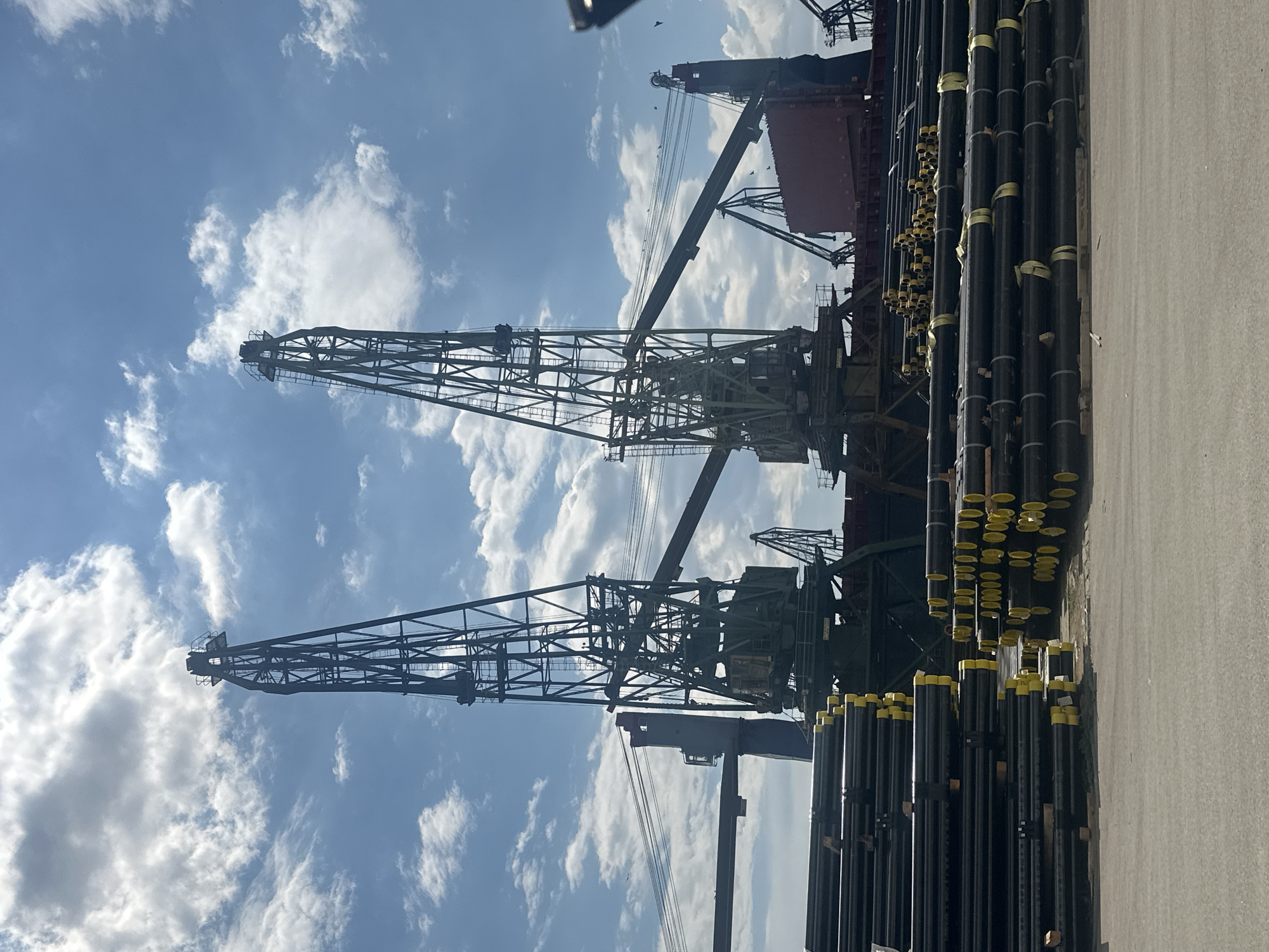 In an outdoor industrial setting, two large green cranes stand prominently against a bright blue sky with scattered white clouds. In the foreground, stacks of black pipes with yellow end caps are neatly arranged on the ground. Behind the pipes, a large red ship is visible, with more cranes and industrial structures in the background. The scene appears to be a port or shipyard, likely for the loading or unloading of materials. The lighting suggests it is daytime, with bright sunlight casting shadows.