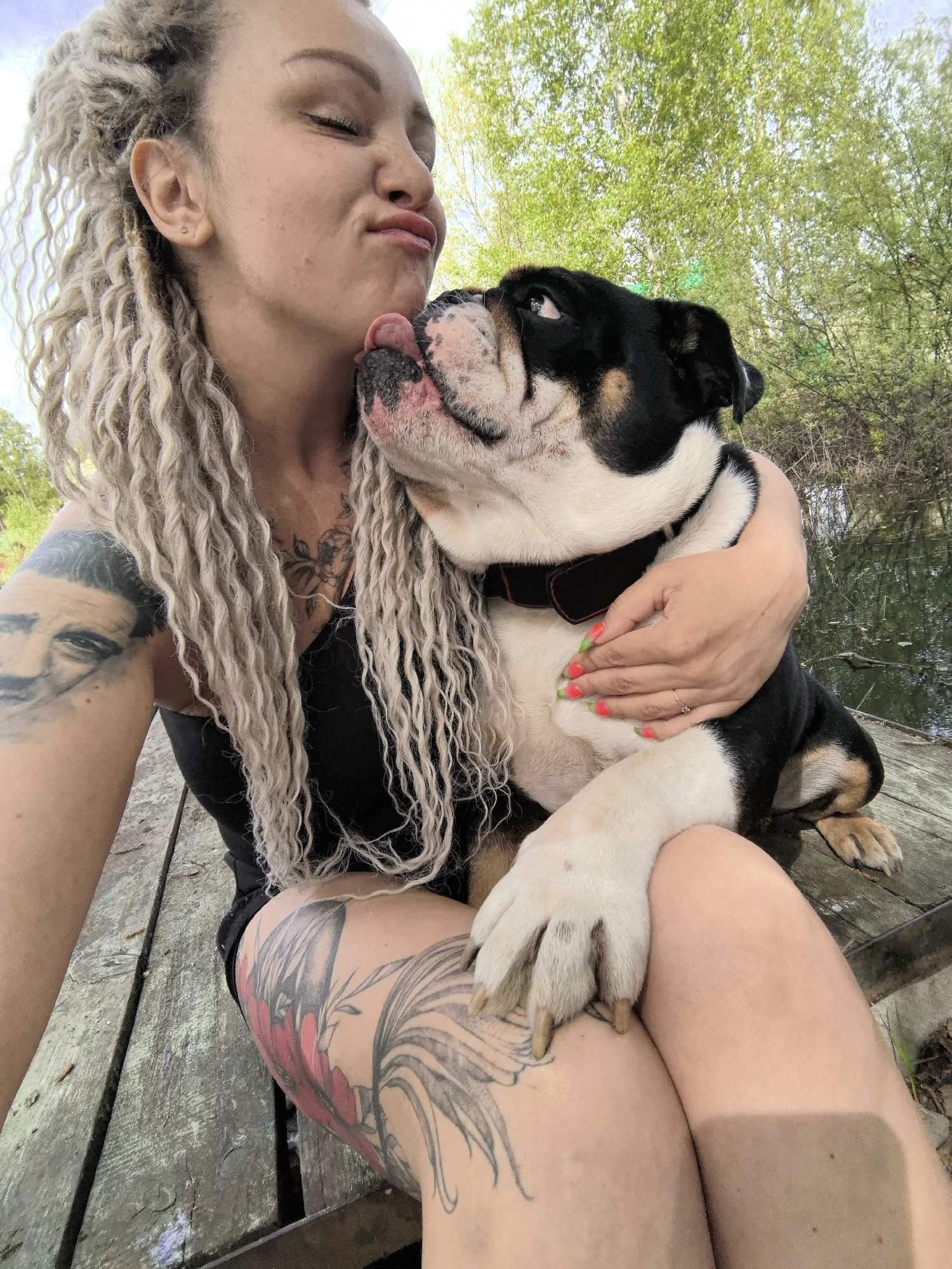 A woman with light blonde dreadlocks and multiple tattoos is seated on a wooden platform outdoors with a large, tri-color bulldog. The woman wears a black tank top and has tattoos on her left arm and thigh. One tattoo on her arm depicts a man's face. The bulldog, primarily white, black, and brown, has its tongue extended towards the woman's face. She is holding the dog in a close embrace. The background includes trees and a body of water. The location is Noviy, Russia.