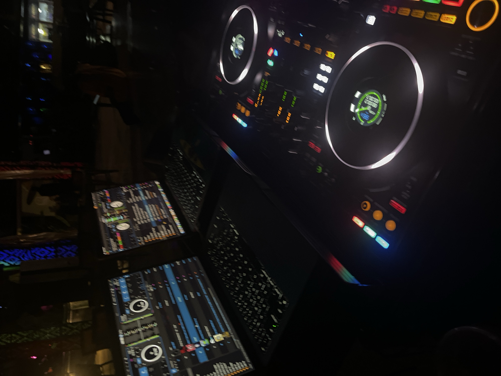 In Howayi, Nigeria, at night, a DJ setup is visible. Two laptops, displaying music software, are positioned on a surface. In the foreground, a DJ controller with illuminated buttons and two turntables is present. The scene appears to be in an outdoor or semi-outdoor area, with dimly lit background elements suggestive of a public space or venue.