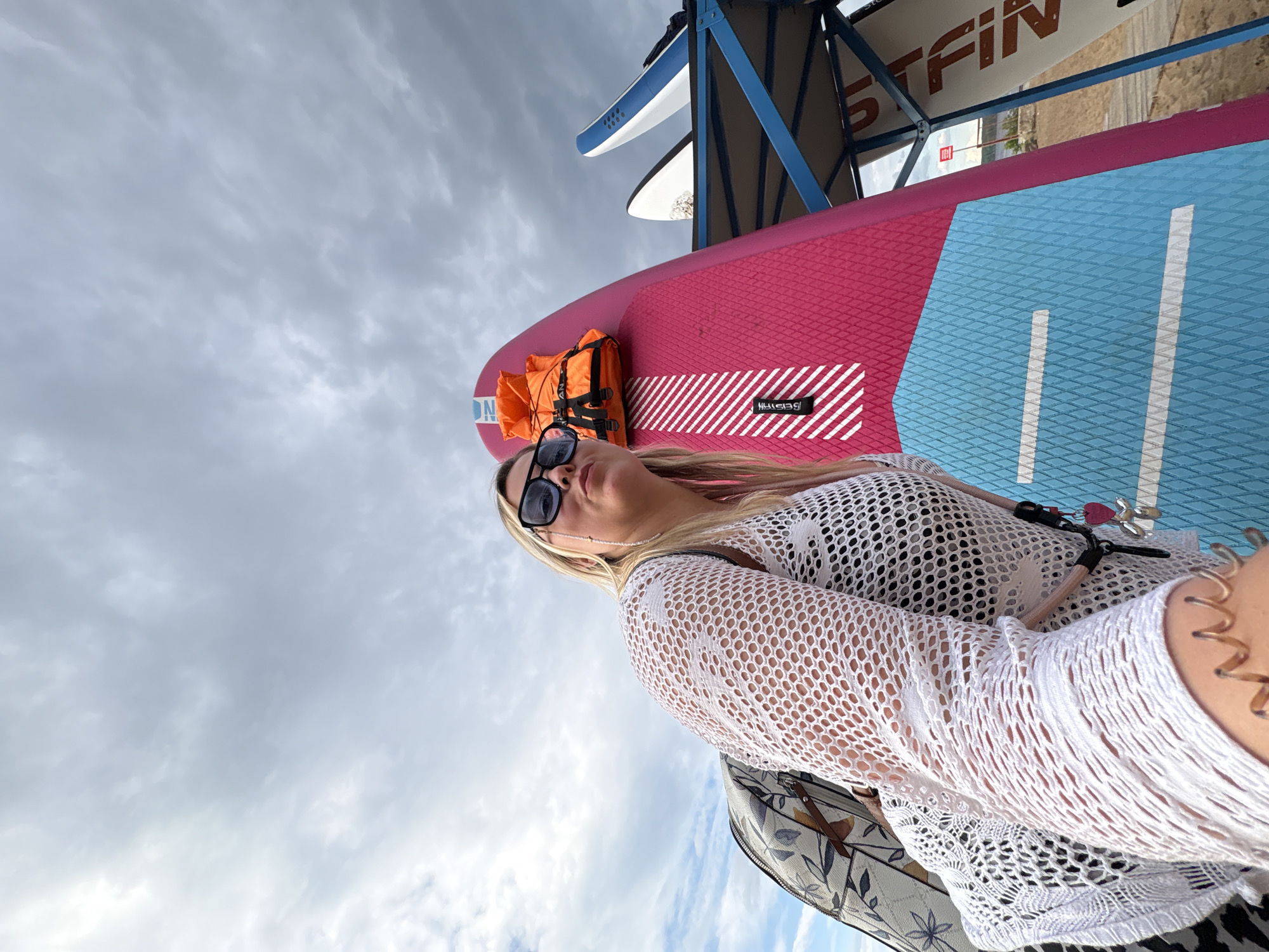 A woman with blonde hair and sunglasses is posing for a photo outdoors. She is wearing a white, open-knit cover-up over a black bikini top and is carrying a backpack with a floral pattern. In the background, there are several surfboards, one of which is pink and blue, and an orange life vest is attached to it. The sky is cloudy. The scene appears to be at a beach or waterfront area, possibly near a place where water sports are available.