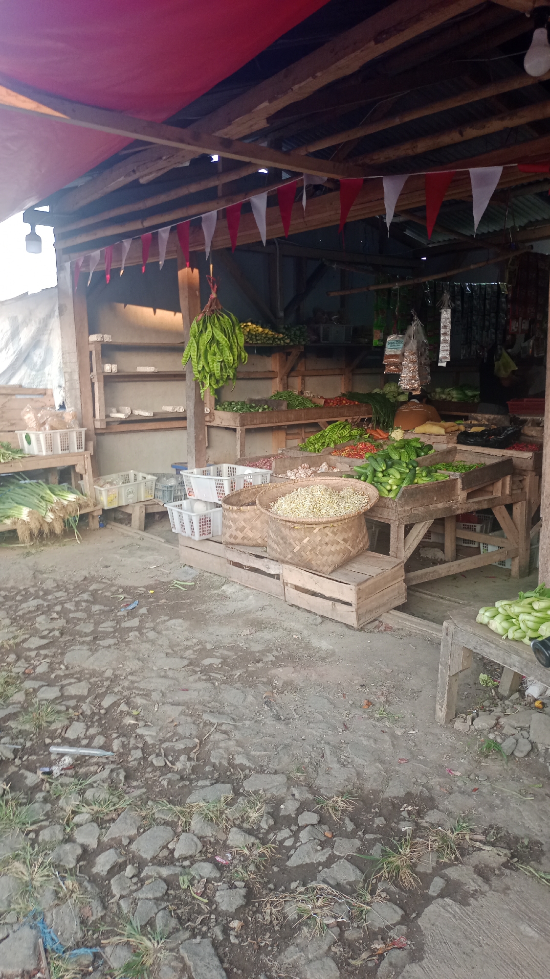 This is a medium shot, exterior, eye-level photograph of an outdoor market stall featuring a variety of fresh produce. The stall is constructed from rough wooden beams and planks, covered by a red tarp roof. Red and white pennant flags are strung across the underside of the roof. In the foreground, the ground is paved with rough, uneven stones and patches of dry grass.
The stall is laden with vegetables. In the center, two large woven baskets are filled with pale yellow sprouts. To the right, a wooden display table is overflowing with cucumbers and other green vegetables, with a pile of red chilies further back. To the left, bundles of green onions are neatly arranged in white plastic crates on a wooden stand. Behind the fresh produce, shelves are visible with some items, and more vegetables are displayed on tiered wooden platforms. A bunch of long, green pods, possibly petai, hangs from the ceiling. The overall impression is of a bustling, rustic market scene. There are no people visible in the photograph. The setting appears to be in Karyamandala, Indonesia, suggested by the presence of local produce and the style of the stall.