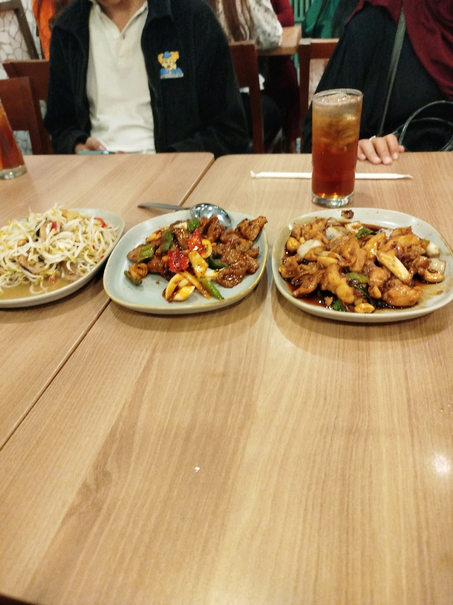 The image captures a close-up, eye-level view of a wooden table laden with food and drinks, likely in a restaurant setting in Balikpapan, Indonesia. The lighting suggests an indoor environment, possibly with ambient restaurant lighting.
In the foreground, on the left, is a plate of stir-fried bean sprouts with some visible pieces of chili and other vegetables, suggesting a side dish or appetizer. To its right is a second plate containing a dish of what appears to be stir-fried meat, possibly beef or chicken, with onions and green bell peppers, coated in a dark, glossy sauce. Further to the right, on a third plate, is another stir-fried dish, also featuring pieces of meat and vegetables in a similar sauce, with onions being a prominent ingredient.
A glass filled with a dark beverage, likely iced tea, sits on the table between the plates, topped with ice cubes. A white straw is visible next to the glass. In the background, portions of people are visible, seated at the table. On the left, a person wearing a black jacket over a light-colored polo shirt is seated, with a portion of their hands visible. On the right, a person wearing a dark headscarf, possibly a hijab, is also seated. The presence of these individuals and the arrangement of food and drinks suggest a shared meal. The wooden table has a visible grain pattern. The overall scene conveys a casual dining experience.