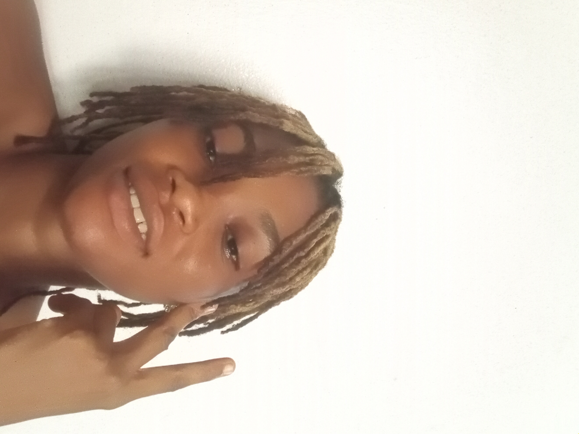 A medium shot of a smiling woman with dreadlocks. Her head is tilted to the left, and her right hand is raised near her face with her index and middle finger extended. The background is a plain white wall. There is no indication of the location being Shagamu, Nigeria.