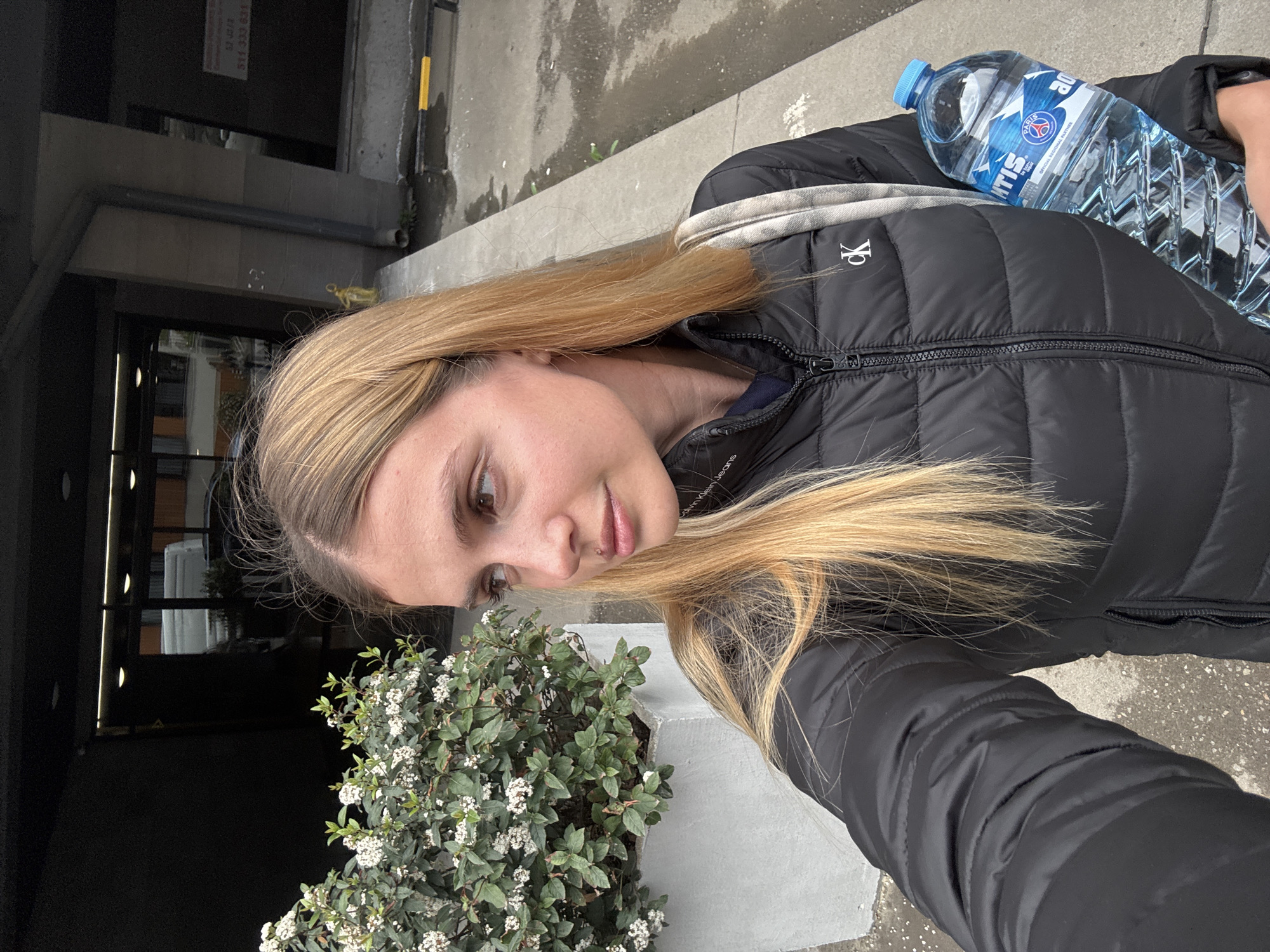 A medium shot captures a fair-skinned woman with long blonde hair, wearing a black Calvin Klein puffer jacket and holding a bottle of water. She is positioned next to a bush with small white flowers, seemingly on a city sidewalk. A building entrance is visible in the background.