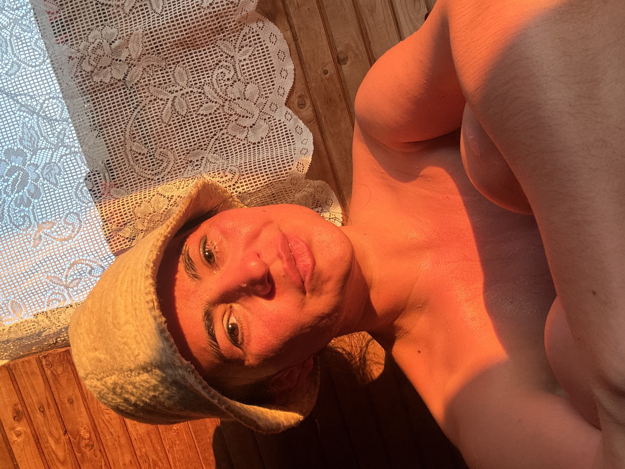 An adult female is depicted in an indoor setting, likely a sauna or traditional Russian banya, indicated by the wooden paneling and the characteristic felt sauna hat she is wearing. She is positioned with her head and upper torso visible, looking directly at the viewer with a calm and relaxed expression, her lips slightly parted in a faint smile.
The scene is bathed in warm, golden light, suggesting either a late afternoon or early morning sunbeam streaming through a window. This strong, directional light illuminates the left side of her face and upper body, casting an orange-red glow on her skin and creating distinct shadows. Her skin appears slightly dewy, consistent with a warm environment.
To her left, a white, intricately patterned lace curtain partially covers a window, allowing the warm light to filter in and create dappled patterns. The background features rustic wooden planks, forming either a wall or a bench, reinforcing the traditional bathhouse ambiance. The overall atmosphere conveyed is one of relaxation and warmth. There is no visible text in the image. The location context provided is Кемуль, Russia, further supporting the interpretation of a banya setting.