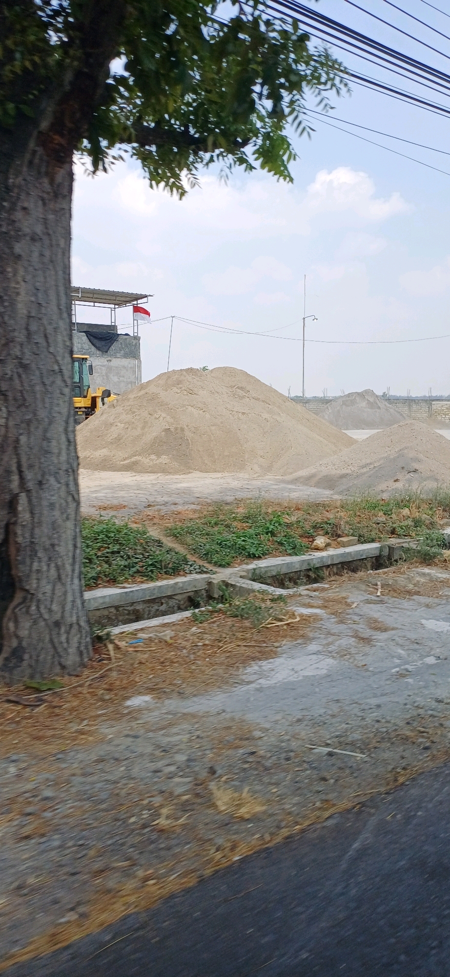 An outdoor scene in Jakenan, Indonesia, shows a construction site with large piles of sand. A yellow bulldozer is partially visible to the left of the main sand pile, parked near a structure with a tin roof and what appears to be a building with a facade of stone-like material. A red and white flag, likely the Indonesian flag, is flying from a pole on top of this structure. In the foreground, a large tree trunk is on the left side of the image, and a drainage ditch runs through the area. Power lines are visible overhead. The ground is a mix of asphalt, gravel, and dry grass. The sky is partly cloudy.