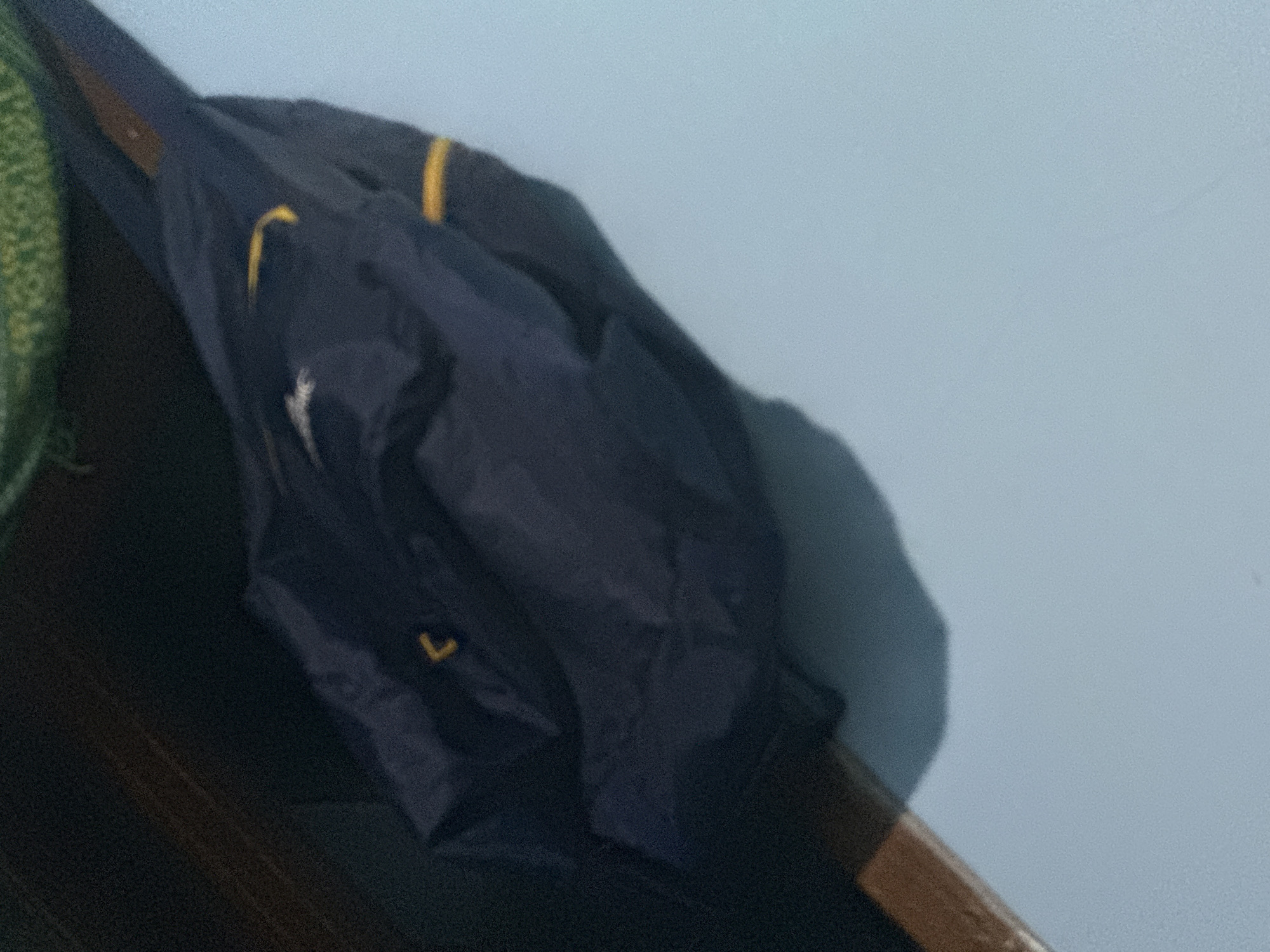 A dark navy jacket with yellow accents is draped over a wooden beam. The jacket appears to be made of a synthetic material and has a slightly reflective sheen. The yellow accents are thin stripes on the sleeves. The background is a light blue wall and a dark, out-of-focus object. The image appears to be taken indoors, possibly in a residential setting in Slahung, Indonesia, as suggested by the context provided. The lighting is dim, casting a shadow of the jacket on the wall.