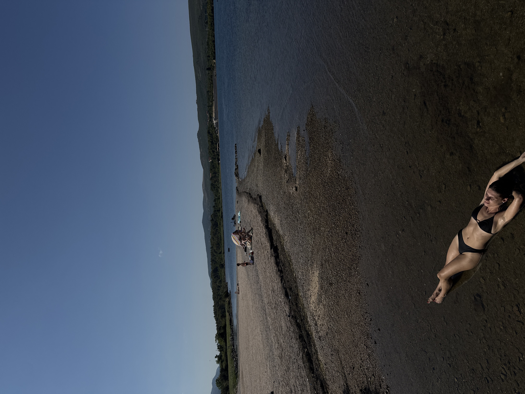 A woman in a black bikini is lying on her back in shallow, clear water at the edge of a stony beach in Mrčevac, Montenegro. Her arms are extended above her head and her eyes are closed. The water is dark and reflects the sky. The beach is composed of small, dark stones and pebbles, with some scattered seaweed. In the background, several people are visible sitting under a beach umbrella, some in swimwear. Further in the distance, there is a body of water with a few boats, surrounded by lush green hills and mountains under a clear blue sky. The sun is shining brightly, casting shadows.