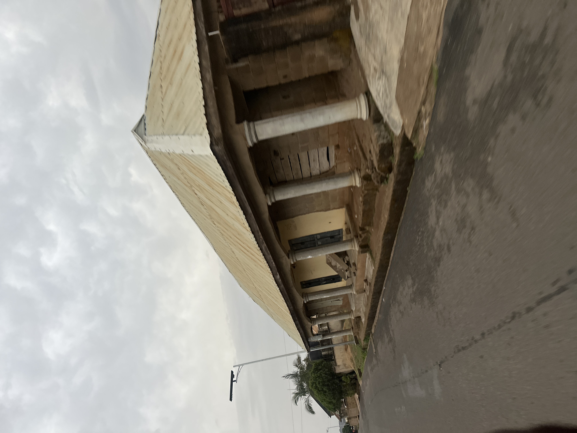 The image depicts a long, single-story building with a colonnaded facade, likely located in Jos, Nigeria. The building is constructed with a corrugated metal roof, brick walls, and cylindrical columns supporting a covered walkway. Some sections of the walkway are boarded up. The building appears old and somewhat dilapidated. A paved road runs along the side of the building. A streetlight pole and a few trees are visible in the background. The sky is overcast. There are no people visible in the frame.