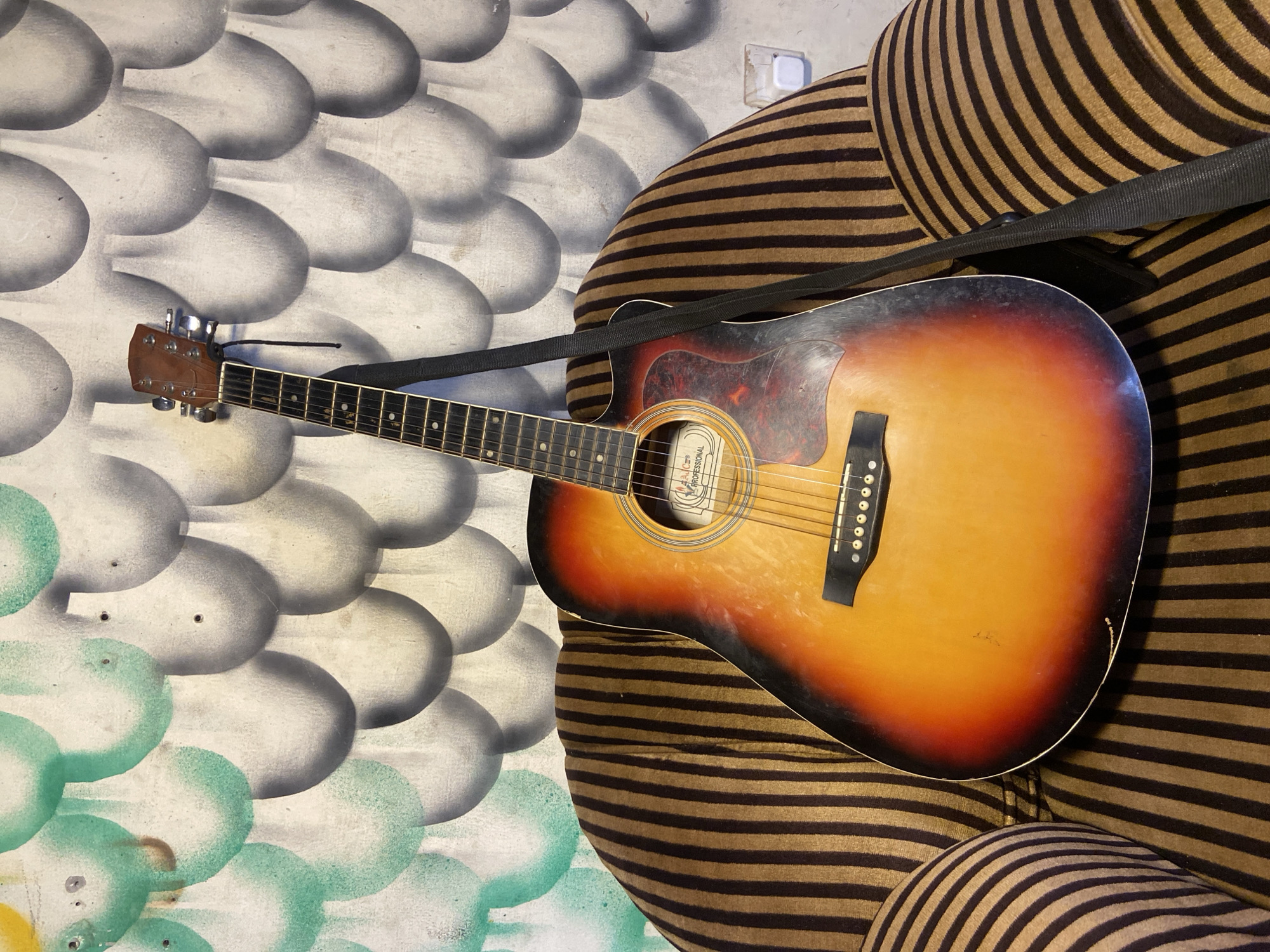An acoustic guitar, featuring a classic sunburst finish with a black pickguard, is depicted leaning against a piece of striped furniture in an indoor setting. The guitar's body transitions from dark edges to a vibrant orange-yellow center, and its headstock, frets, and six strings are clearly visible. A black strap is attached to the guitar and draped over the furniture. Minor wear and dust are visible on the instrument.
The furniture, likely a chair or sofa, is covered in a fabric with distinct horizontal black and brown stripes. Behind the guitar and furniture, a wall is adorned with a painted pattern resembling overlapping oval or scale-like shapes. The upper portion of this wall pattern is rendered in grey and white tones, while the lower left section incorporates similar shapes in shades of green. An electrical outlet is visible on the wall above and to the right of the furniture.
Inside the guitar's soundhole, a label is partially visible, displaying the word "PROFESSIONAL". The scene suggests a casual, lived-in environment in Mararaba, Nigeria. No individuals are present, and the lighting indicates an indoor setting.