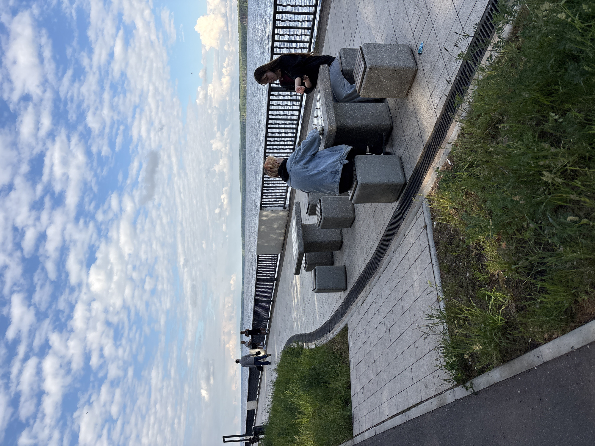 The image depicts a waterside promenade or embankment, likely along a large river or lake, under a bright, cloudy sky. The scene is oriented vertically in the original image but described here as if correctly rotated.
In the mid-ground, two individuals are seated at a gray concrete bench that incorporates a game board, possibly for chess or checkers. One person, with light-colored hair, wears a blue denim jacket and sits with their back partially to the camera, facing the board. Opposite them is a person with long, dark hair, wearing a black top and light-colored pants, actively engaged with the game board. Further along the promenade, two more people are visible near the dark metal railing that separates the paved area from the water.
The promenade features light gray rectangular pavers and a long, dark drainage grate running parallel to the water. Beyond the grate and a curb, there is a strip of wild green grass and weeds, bordering a darker asphalt path on the far right. In the background, a wide expanse of water stretches towards a distant tree-lined shore.
The sky is a prominent feature, largely blue with numerous scattered, fluffy white clouds, indicating fair weather during daytime. The lighting suggests late morning or afternoon. A small, blue object, possibly a cap or bottle, is visible on the paved ground near one of the benches. There is no visible text in the image.