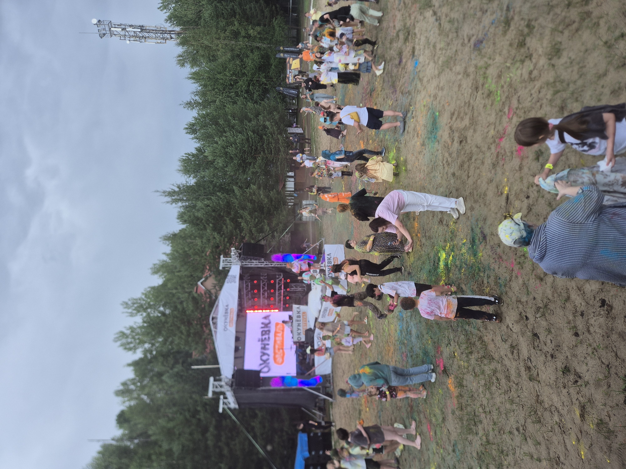 An outdoor event, likely a festival, is taking place with a crowd of people gathered around a stage. The ground is sandy and covered with colorful powder, suggesting a Holi-like celebration. People of various ages are present, many are dressed casually, and some have powder on their clothes and skin. The stage has a large screen displaying text and logos, with lights and speakers set up. In the background, there are trees and a cellular tower. The overall atmosphere appears festive and energetic, with people dancing and interacting.