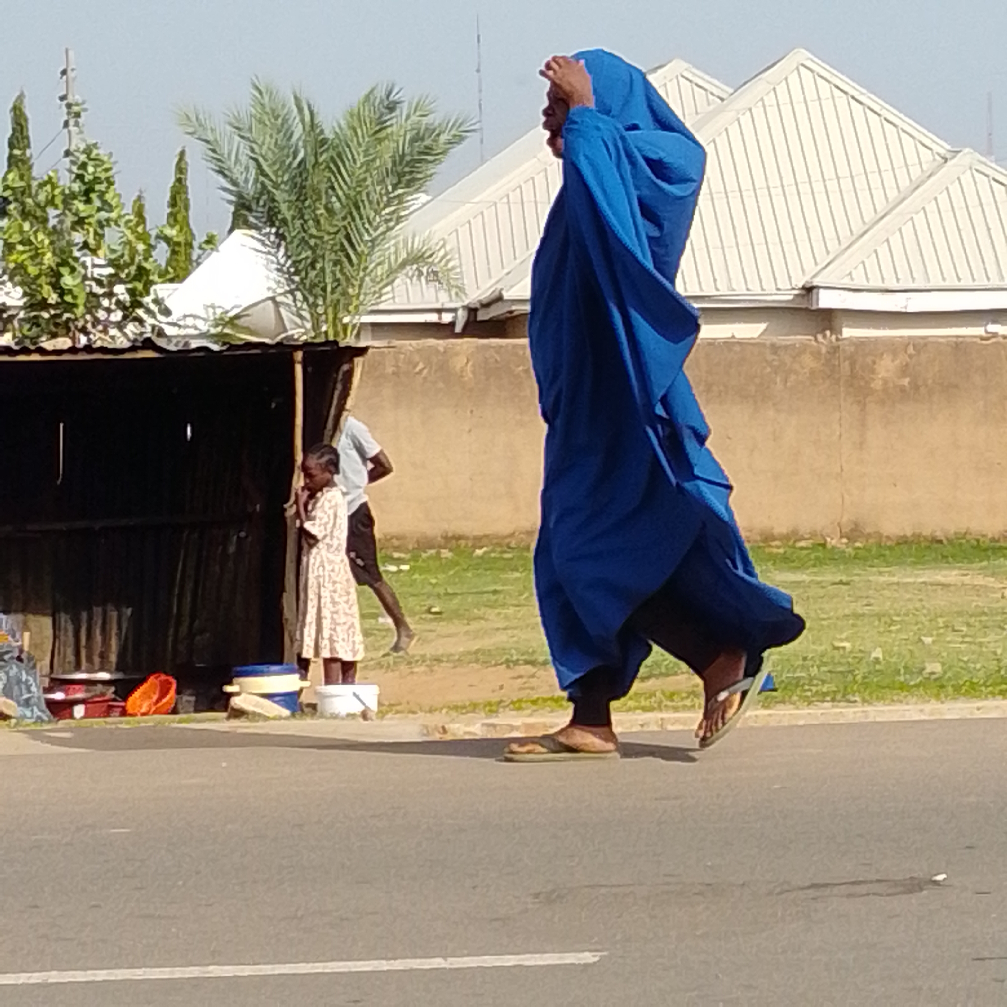 The image captures an outdoor scene on a street in Tempure, Nigeria, under a clear sky. In the foreground, a person draped in a vibrant blue garment is walking, their feet clad in sandals. To the left, a small roadside stall with a dark, corrugated iron roof is visible. A young girl in a patterned dress stands near the stall, looking down. Behind her, a man in a white t-shirt and dark shorts walks away, his posture suggesting movement. The environment includes a dirt and grass area bordering the street, with a light-colored wall and buildings with white, sloped roofs in the background. Several trees, including a palm tree, add to the natural elements of the scene. Objects on the ground near the stall include various containers and what appear to be plastic items. The overall impression is of a typical street scene in a Nigerian urban or semi-urban setting.