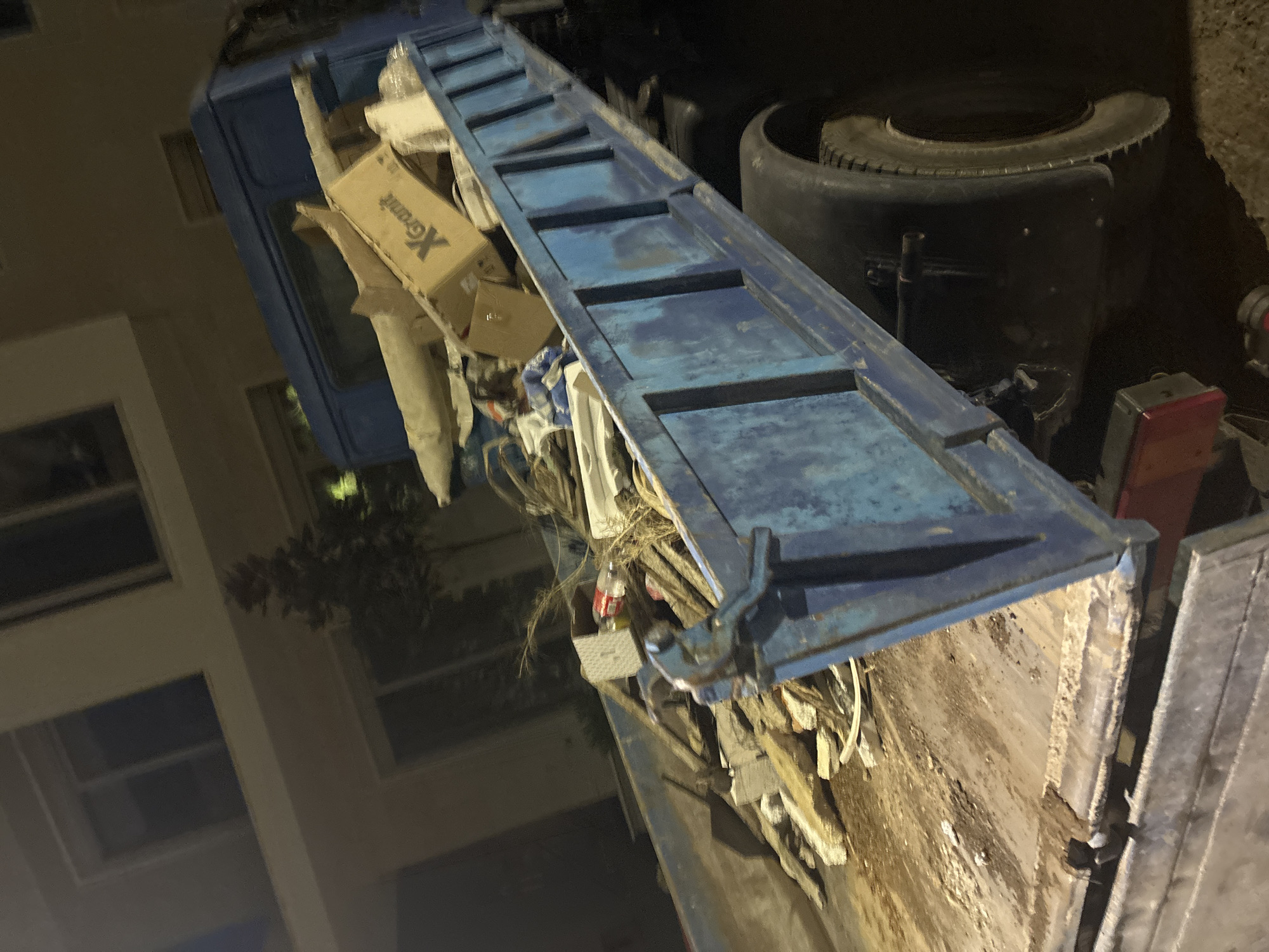 The image shows a close-up view of a blue garbage truck filled with debris, including cardboard boxes, plastic, and organic waste. The truck appears to be in a residential or urban setting, with a building visible in the background on the left. The lighting suggests it might be nighttime or early morning.
The most prominent object is the truck's blue metal bed, which is loaded with a jumble of waste. A partially visible cardboard box with the text "Xeramit" printed on it is noticeable among the refuse. Also, a spare tire is stacked on top of the truck's chassis.
The scene is cluttered and disorganized, characteristic of a garbage collection operation. The angle of the photograph, tilted to the right, gives a dynamic perspective of the scene. The surrounding environment is dark, indicating a lack of strong natural light. The building in the background has multiple windows, suggesting it is an apartment building. There are no people visible in the image, and the focus is entirely on the truck and its contents. The overall impression is that of a functional, albeit somewhat unglamorous, task being carried out in a semi-urban environment at night.