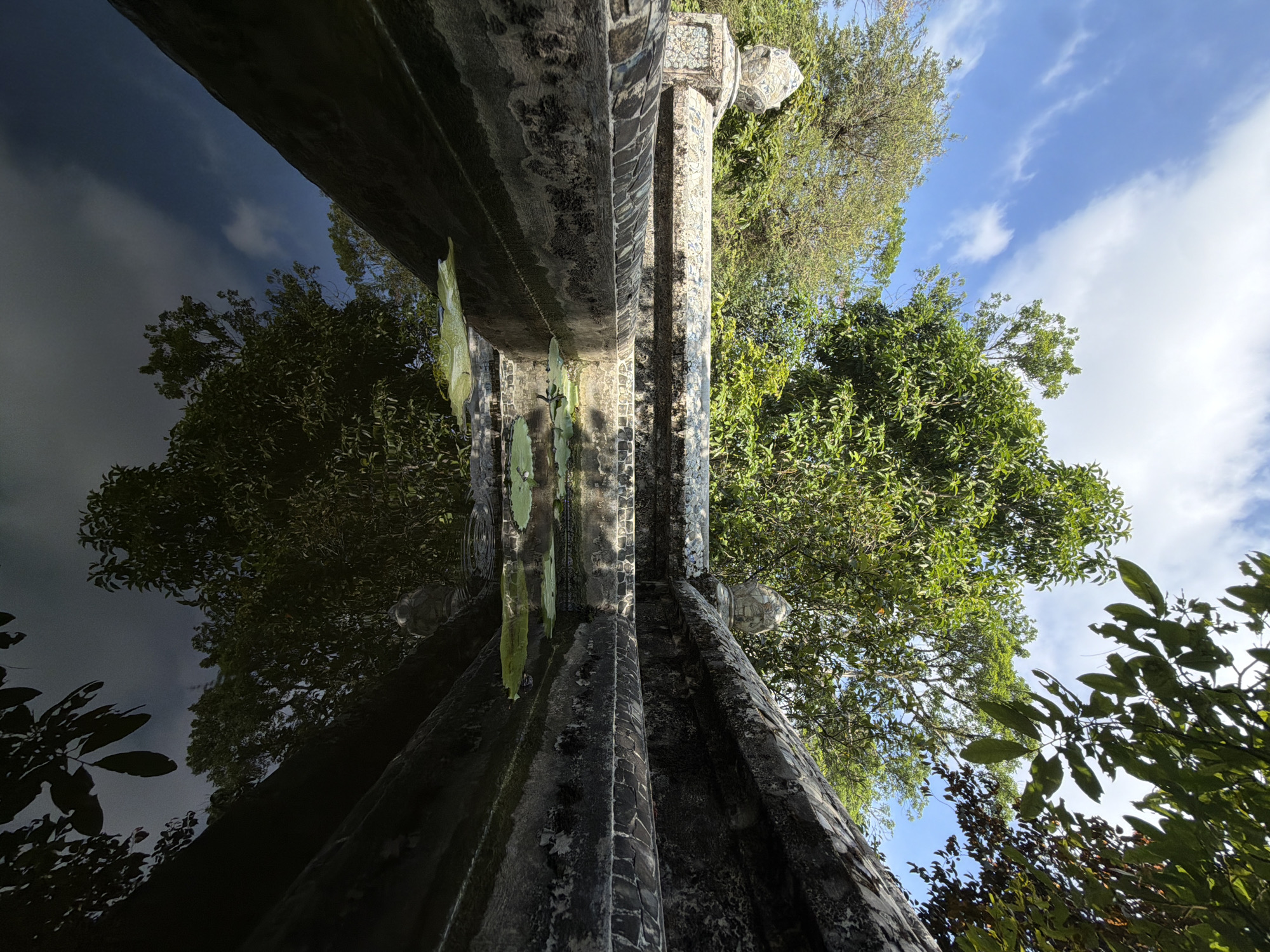 A low-angle perspective captures a detailed architectural structure, possibly a bridge or decorative wall, made of weathered concrete or stone. The surface is textured with what appears to be decaying tiles and some plant growth. The structure intersects with a reflective body of water, mirroring the trees and sky above. The sky is partially clear with scattered clouds. The location is Thủy Xuân Ward, Vietnam.